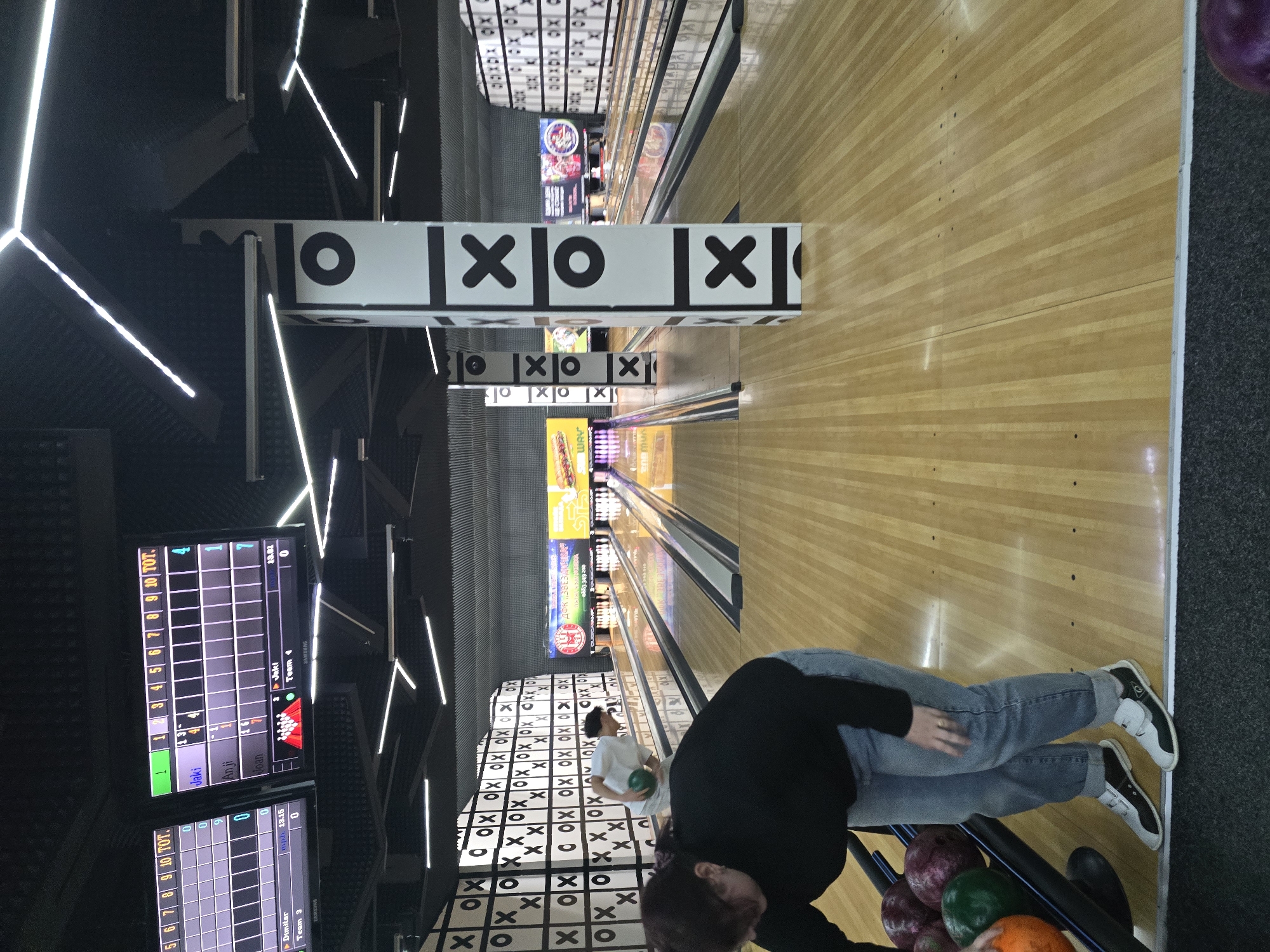 The image shows the interior of a bowling alley. Lanes stretch into the distance under overhead tic-tac-toe decor. A scoring screen is visible displaying the scores for Jaki, Anji, and Joan in Team 4 and Dimitar in Team 3. A person, presumably about to bowl, stands near a rack of bowling balls, with an orange ball in view. The floor leading to the lanes has a tic-tac-toe pattern. Advertisements for food items line the upper lanes. A white-shirted person stands between the lanes. The image was possibly taken in Burgas, Bulgaria, given the presence of the Cyrillic text "ЗВЕЗДИЧКА" and a Bulgarian phone number, "0883349210."