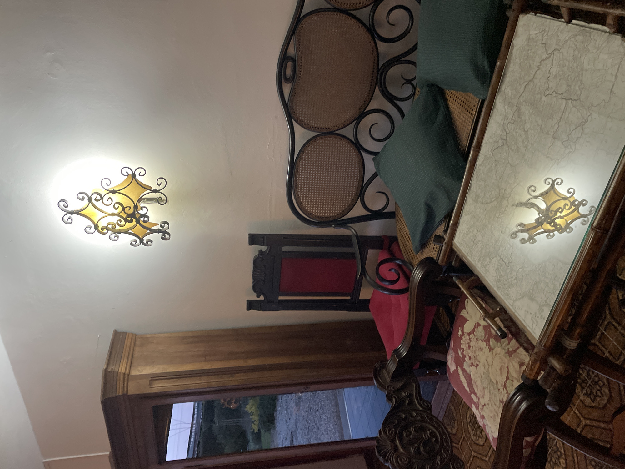 The photograph depicts an interior scene with antique furniture and lighting. A wall-mounted ornate light fixture casts a glow on the adjacent white wall. Visible furniture includes a caned settee with dark green pillows, an antique mirror or table with a reflective surface reflecting the light fixture, and a chair with patterned upholstery. A door or window is visible in the background, looking out onto what appears to be a street or outdoor area. While the furniture style suggests a vintage or classic Italian aesthetic, the location in San Lorenzo, Italy, is not definitively verifiable from the visual information provided. The scene conveys a sense of historical or traditional Italian interior design.