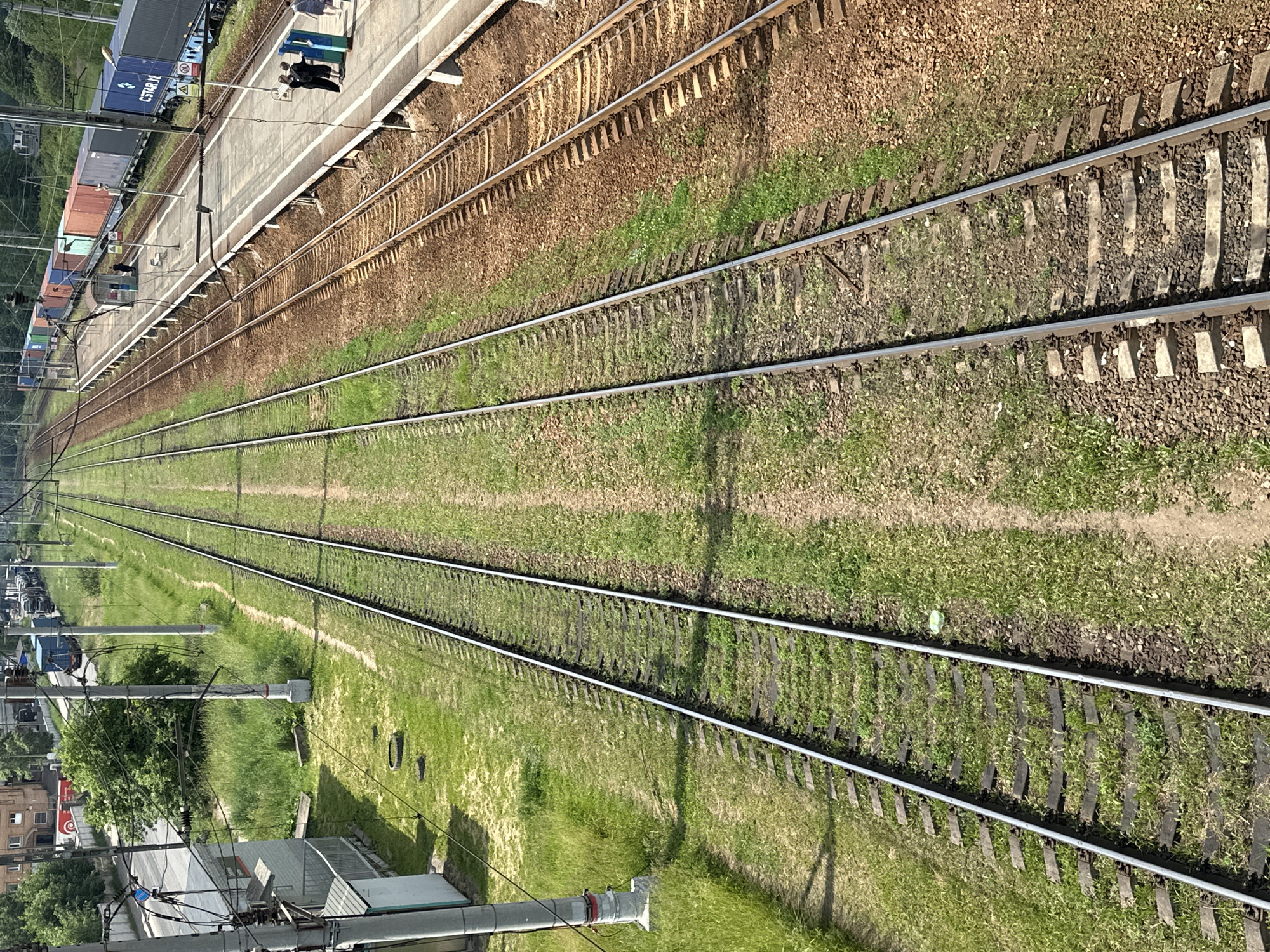 An aerial perspective captures multiple train tracks at a railway station in Selyatino, Russia. The foreground features two sets of parallel train tracks, one on the left flanked by green grass and the other on the right bordered by brown ballast and a concrete platform. In the midground, several cargo train cars, including shipping containers of various colors, are parked on the tracks along the platform. A small blue and gray structure, possibly a signal box or waiting shelter, is situated on the platform. In the background, more train cars and tracks are visible, along with overhead electrical wires and poles, and some buildings and trees lining the railway. Two individuals in dark clothing are standing on the platform, and another person in a blue shirt stands further down the platform. A few more individuals are visible further in the background on the platform. The scene is illuminated by daylight, casting shadows on the tracks and platform.