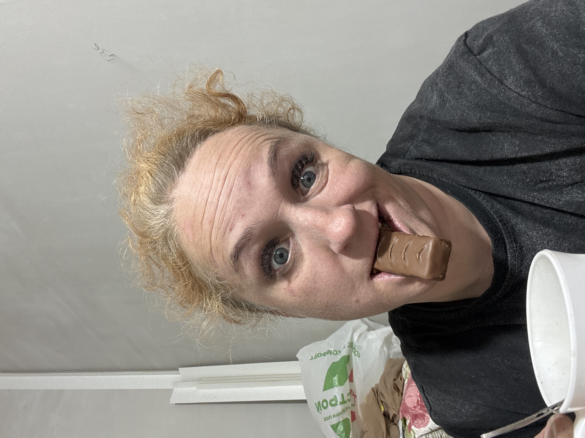 A woman is holding a chocolate bar in her mouth and looking at the camera. She has light blonde curly hair, fair skin, and blue eyes. She is wearing a dark gray t-shirt. In the bottom right corner of the image, there is a white mug and a metal spoon. There is a white plastic grocery bag with green text on it visible behind the woman. The image appears to have been taken inside a building with a white ceiling and white trim. The grocery bag has Russian writing, suggesting the image was taken in Russia. Given the word "Строй" is visible on the bag, this is likely a "Stroykomfort" store. There is a store of that name in Chaikovsky, Russia.