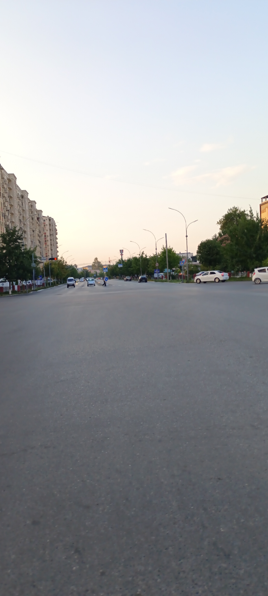 The image depicts a wide, multi-lane asphalt road in what appears to be a city, with apartment buildings lining the left side and trees and streetlights on both sides. The sky is a pale, clear blue, suggesting it is daytime, likely late afternoon or early evening due to the soft lighting. Several cars are visible on the road, indicating traffic. A pedestrian crossing with a person in the middle of it is present, suggesting a functional intersection. Streetlights and traffic lights are also visible, common urban features. The architecture of the apartment buildings suggests a modern urban environment. While no specific text is clearly legible, the presence of road signs implies a developed infrastructure. The overall impression is a snapshot of daily urban life in a city like Fergana, Uzbekistan, during a calm period with moderate traffic.