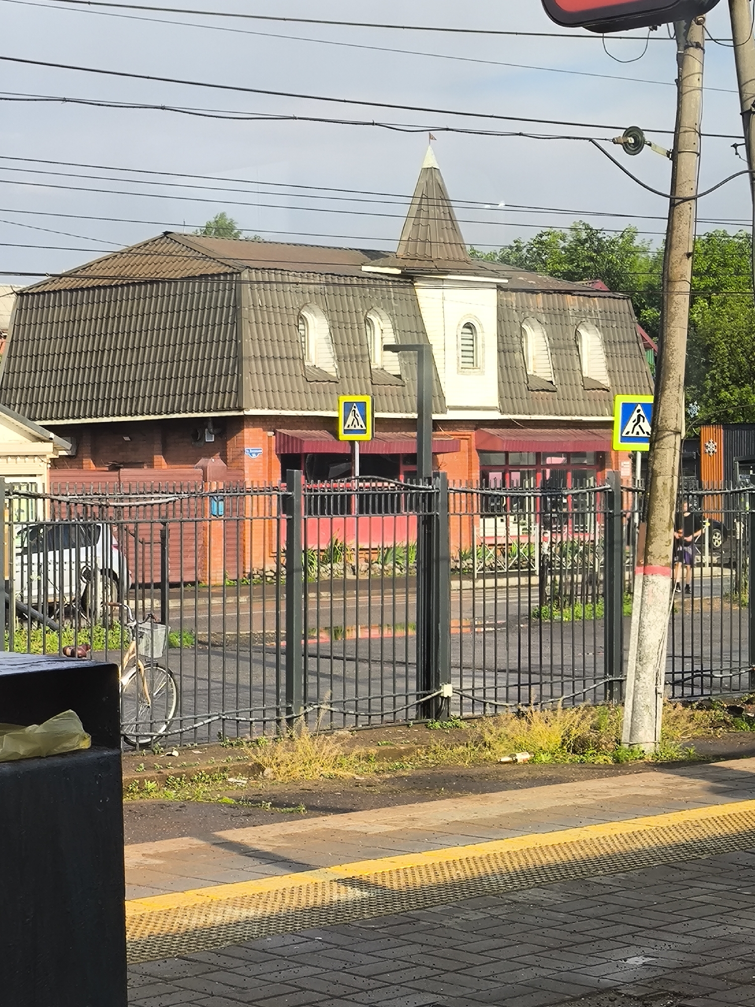 The image shows a street scene in Lobnya, Russia, with a prominent building featuring a distinctive Mansard roof and a small turret. The building is constructed with red brick on the lower level and has arched windows. A black metal fence runs across the foreground, separating the street from the building's property. Several yellow pedestrian crossing signs are visible, indicating a public thoroughfare. A bicycle with a basket is parked near the fence. A black trash bin is on the left side of the image, with a yellow plastic bag inside. In the background, trees and utility poles with overhead wires are present. A person wearing dark clothing and shorts is walking on the sidewalk in the right portion of the image. The ground is paved with interlocking bricks and has a yellow tactile warning strip along the edge.