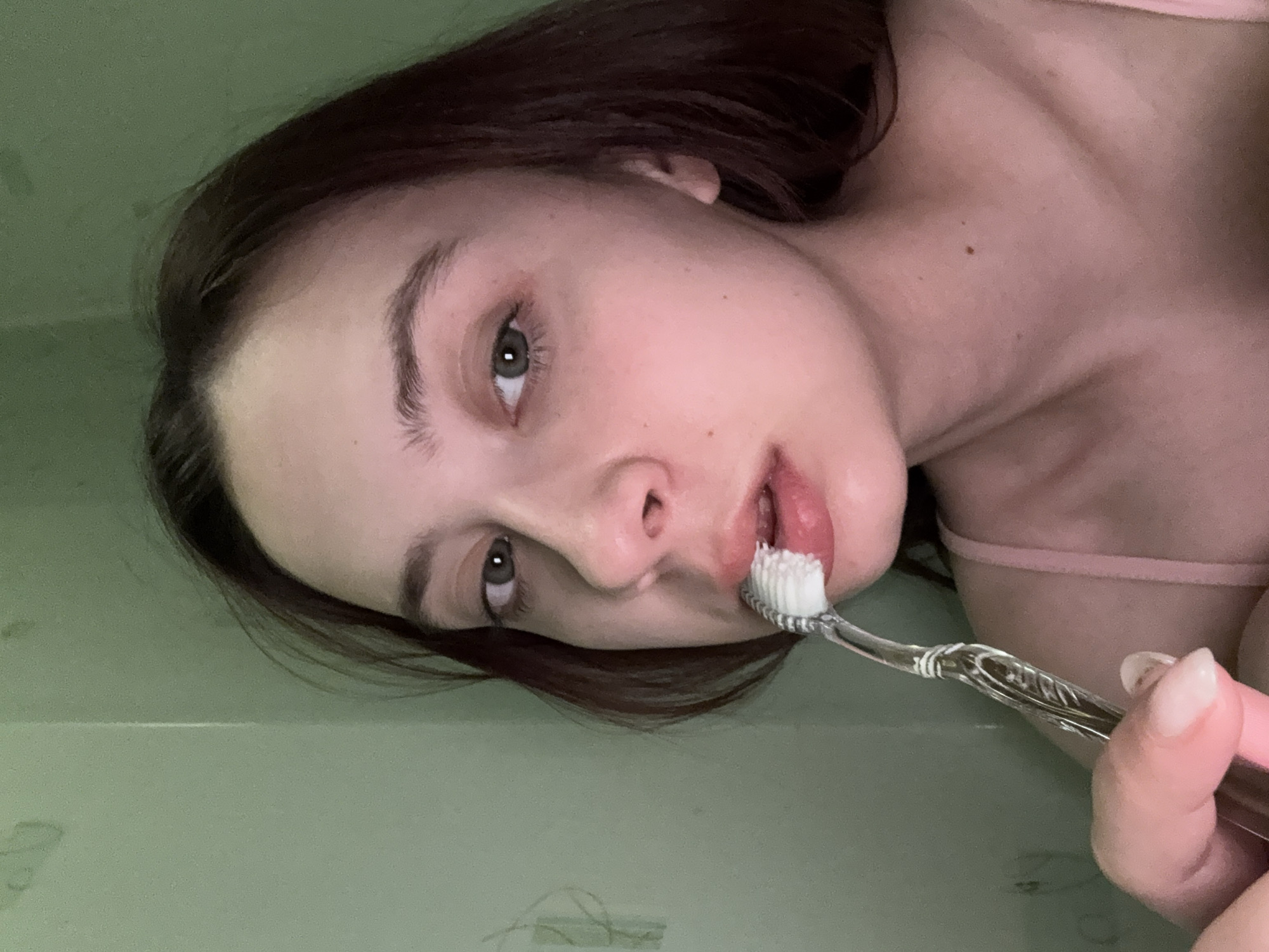 A woman with light skin, brown hair, and green eyes is holding a toothbrush to her mouth. She is wearing a pink top. The background appears to be a bathroom wall with some markings. There is no indication of this image being in the city of Прикамский, Russia.