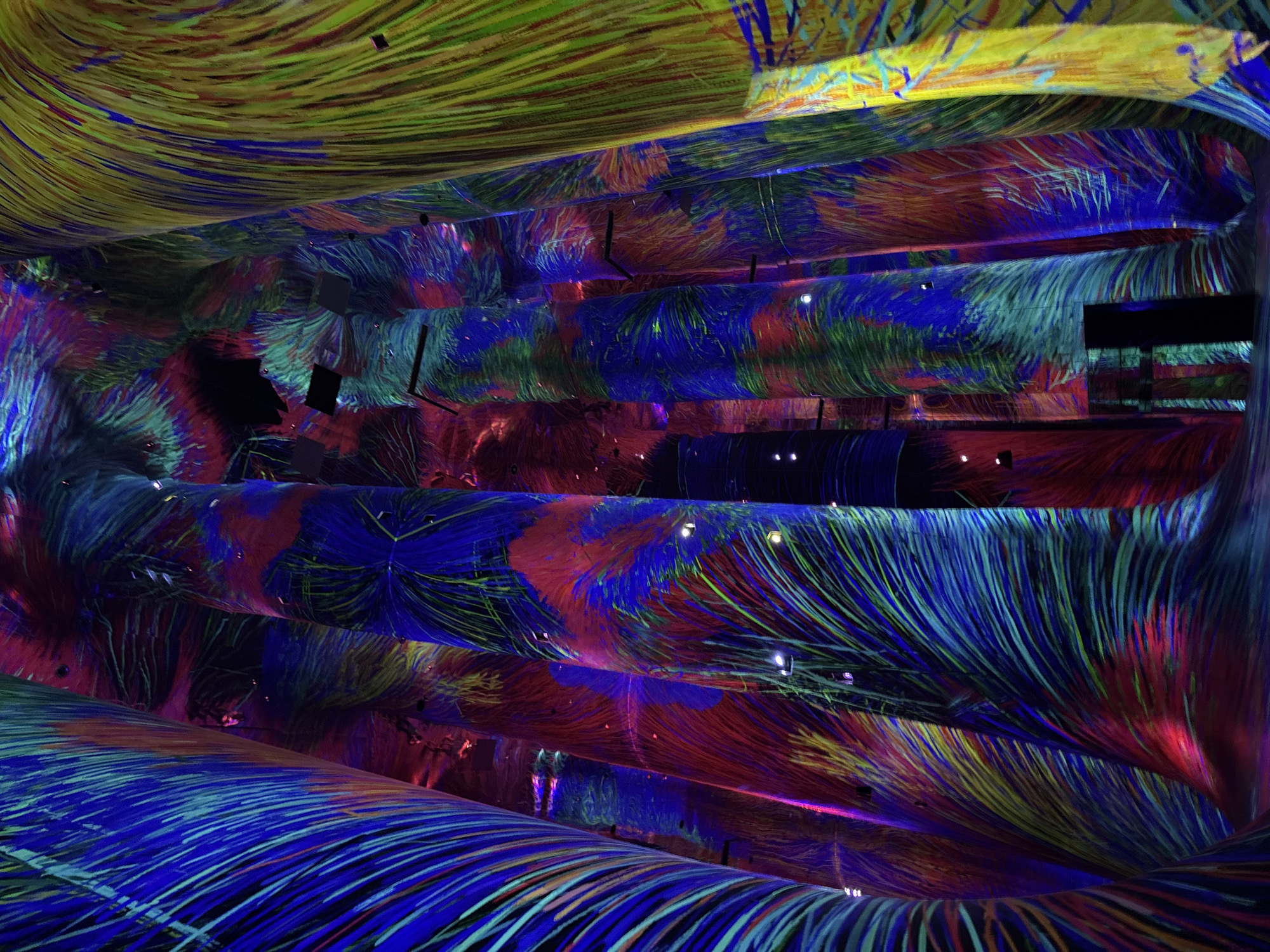 The image depicts the interior of a large, dimly lit space, possibly an exhibition hall or a museum exhibit, in Abu Dhabi, United Arab Emirates. The dominant feature is the projection of vibrant, abstract digital art onto tall, cylindrical structures resembling pillars or columns. These projections create a dynamic and immersive visual experience, with swirling patterns of intense colors like electric blue, deep red, bright yellow, and emerald green. The lines within the projections appear fluid and organic, suggesting movement and energy. Small, square-shaped objects, possibly speakers or lighting fixtures, are intermittently visible against the projected patterns on the columns. The floor is dark, and the ceiling is mostly obscured by the projected light, contributing to the sense of being enveloped by the artwork. The overall impression is one of a technologically advanced and visually stimulating environment designed to engage visitors with contemporary digital art installations.