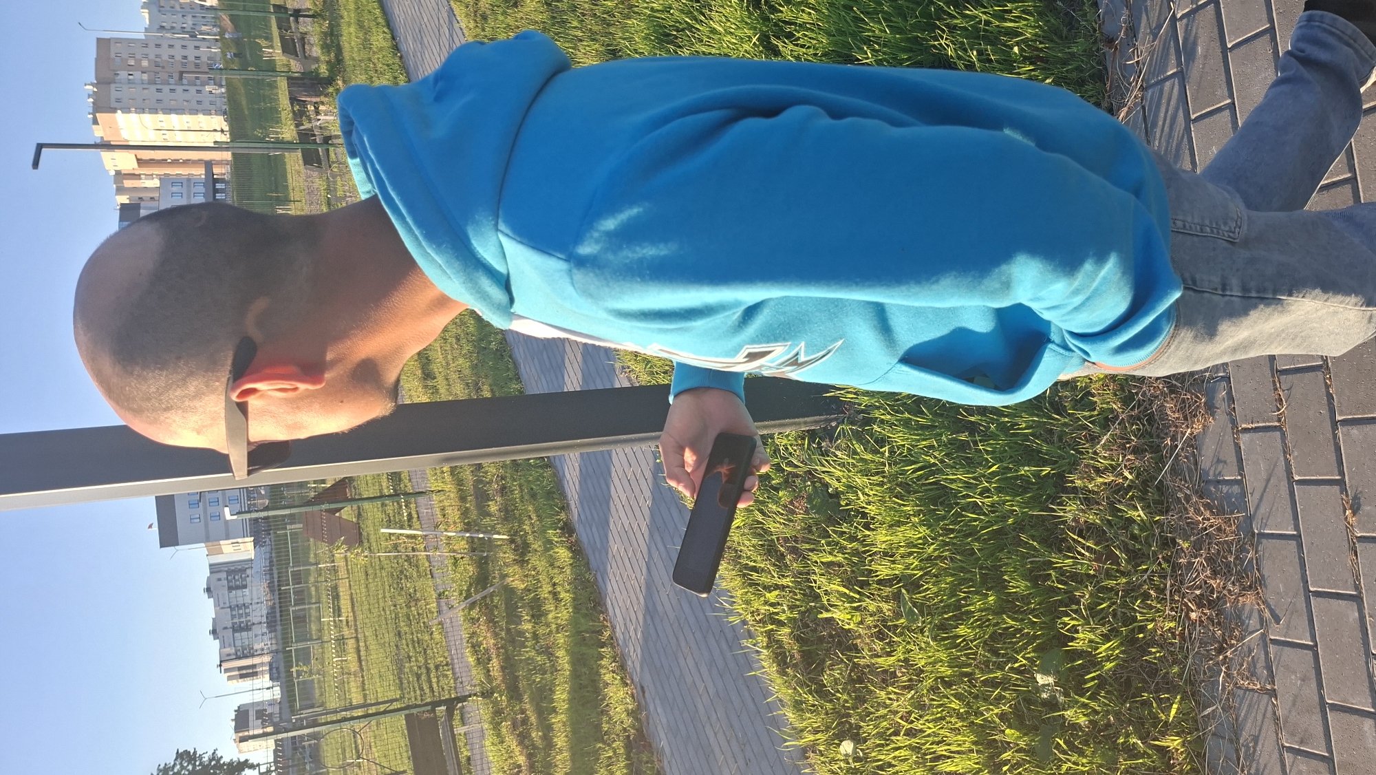 A person with a shaved head and sunglasses is walking on a paved sidewalk next to a grassy area. They are wearing a blue hooded sweatshirt and jeans, and holding a black smartphone in their right hand. In the background, there are apartment buildings and a park with benches and a fence. The scene appears to be in a residential area during daylight. The location is identified as Smaliavichy, Belarus.