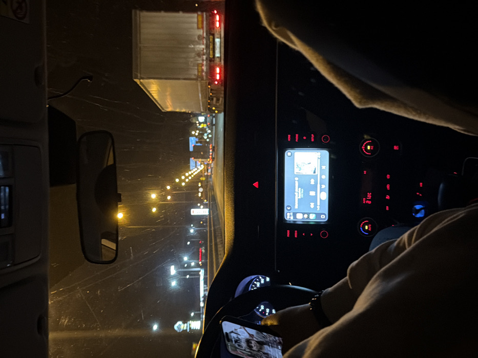 The image captures an interior view from a moving vehicle at night, in Pavlovskaya, Russia, looking forward through the windshield. A person, presumably the driver, has their left hand on the steering wheel and holds a smartphone in their right hand. A black wristband or watch is visible on the left wrist. The smartphone screen displays an image of an individual. The vehicle's central infotainment screen is illuminated, showing a music player interface with the title "Памятный романс" and album art. Below it, the climate control displays "21.0C," and the red triangular hazard light button is illuminated. Outside the vehicle, the road is brightly lit by numerous yellow streetlights extending into the distance. A large white semi-trailer truck with illuminated red taillights is visible in the adjacent lane to the right. Additional vehicle lights are present further down the road, and a lit commercial establishment, possibly a gas station, is visible on the left. A partial view of a passenger's leg, clad in light-colored fabric, is present on the far right of the frame. The windshield exhibits streaks or marks.