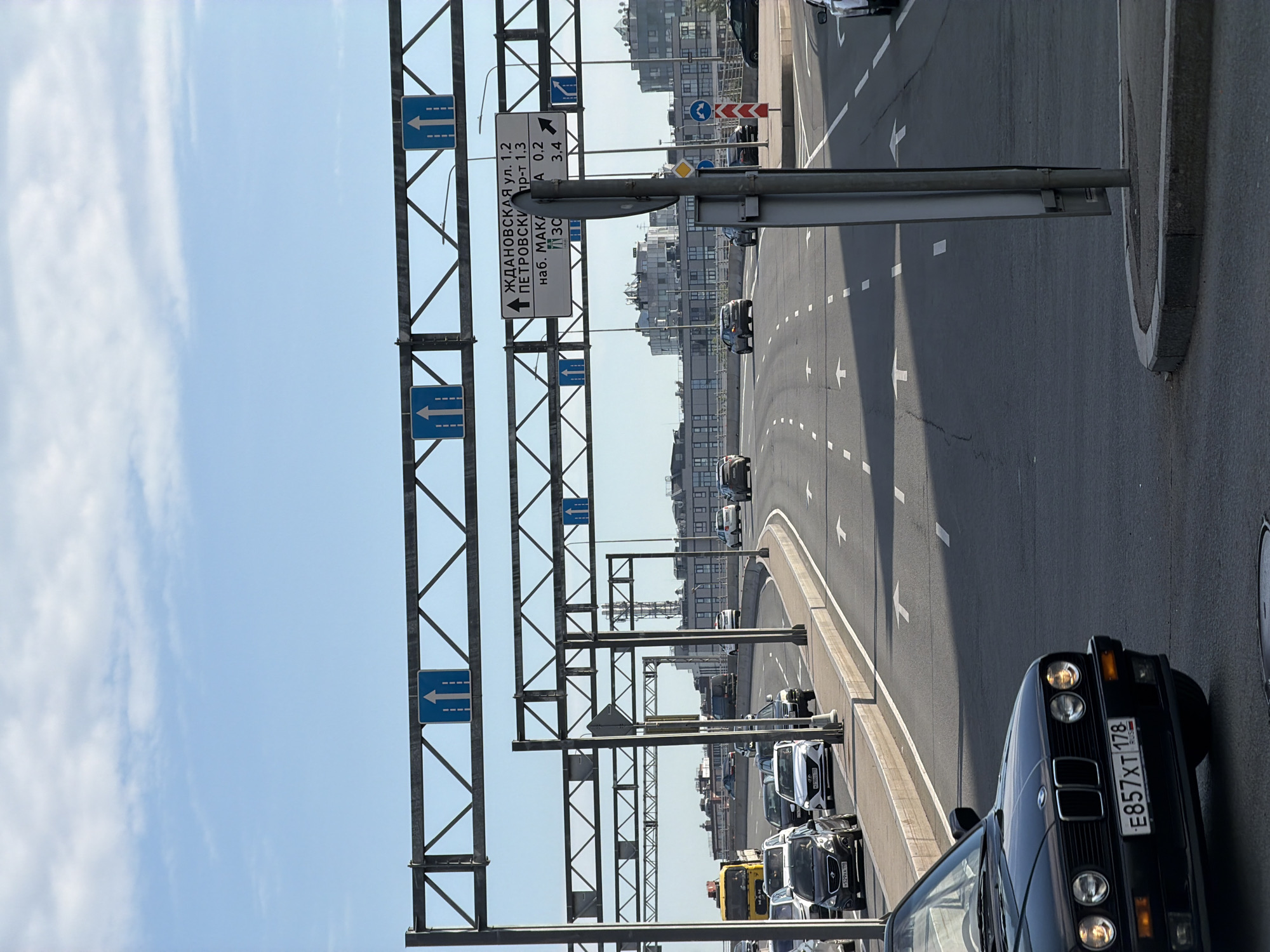 The image shows a multi-lane road in Saint Petersburg, Russia, under a bright blue sky with scattered clouds. Overhead gantries display road signs with directional arrows, indicating straight ahead and right turns. One sign provides distances to various locations, including "ЖДАНОВСКАЯ ул. 1.2", "ПЕТРОВСКИЙ пр-т 1.3", and "наб. МАКАРОВА 0.2" and "пл. 30". The road is marked with white lane lines and arrows. Several cars are visible on the road, including a black BMW in the foreground, a yellow truck, a white sedan, and various other passenger vehicles. The road curves to the right in the distance, leading towards a city skyline with modern buildings and industrial structures. The scene depicts a typical urban traffic environment.