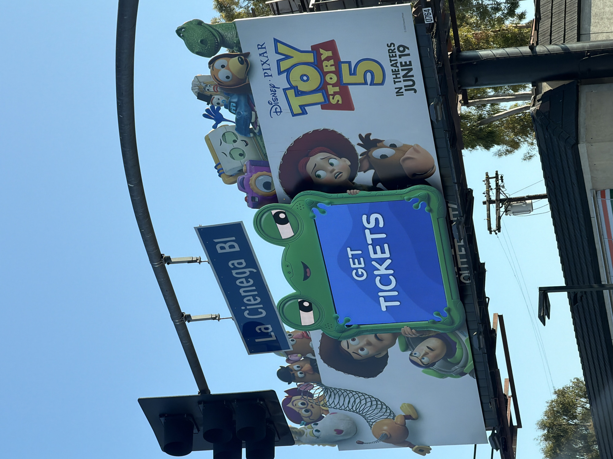 A wide billboard for "Toy Story 5" is positioned above a street. The billboard features characters from the movie, including Woody, Buzz Lightyear, Jessie, Rex, Slinky Dog, and Mr. and Mrs. Potato Head. A large green frog graphic with cartoon eyes is placed in front of the billboard, holding a blue screen that reads "GET TICKETS". The "Toy Story 5" logo is prominent, with the release date "IN THEATERS JUNE 19" displayed below it. A street sign for "La Cienega Bl" is attached to an overhead traffic light pole. The sky is clear and blue. Trees and buildings are visible in the background. The image is taken from a low angle, suggesting the viewer is in a vehicle.