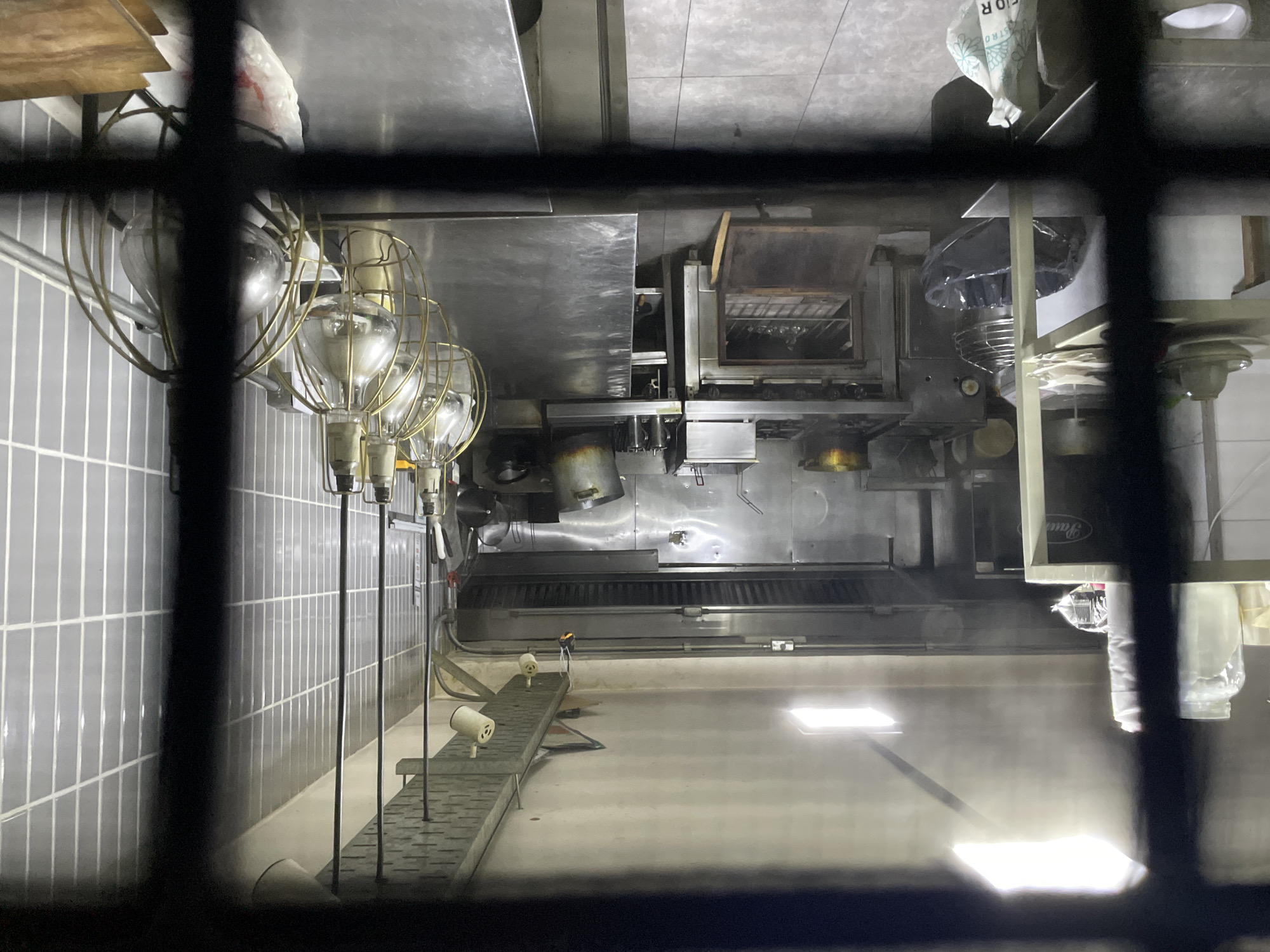 A photograph taken through a window shows the interior of a commercial kitchen. Stainless steel counters and appliances, including an oven, are visible. A row of industrial-style lights hangs along a gray tiled wall. The floor is light-colored. There is no evidence of people, specific subjects, actions, or interactions visible in the scene. The location is uncertain.