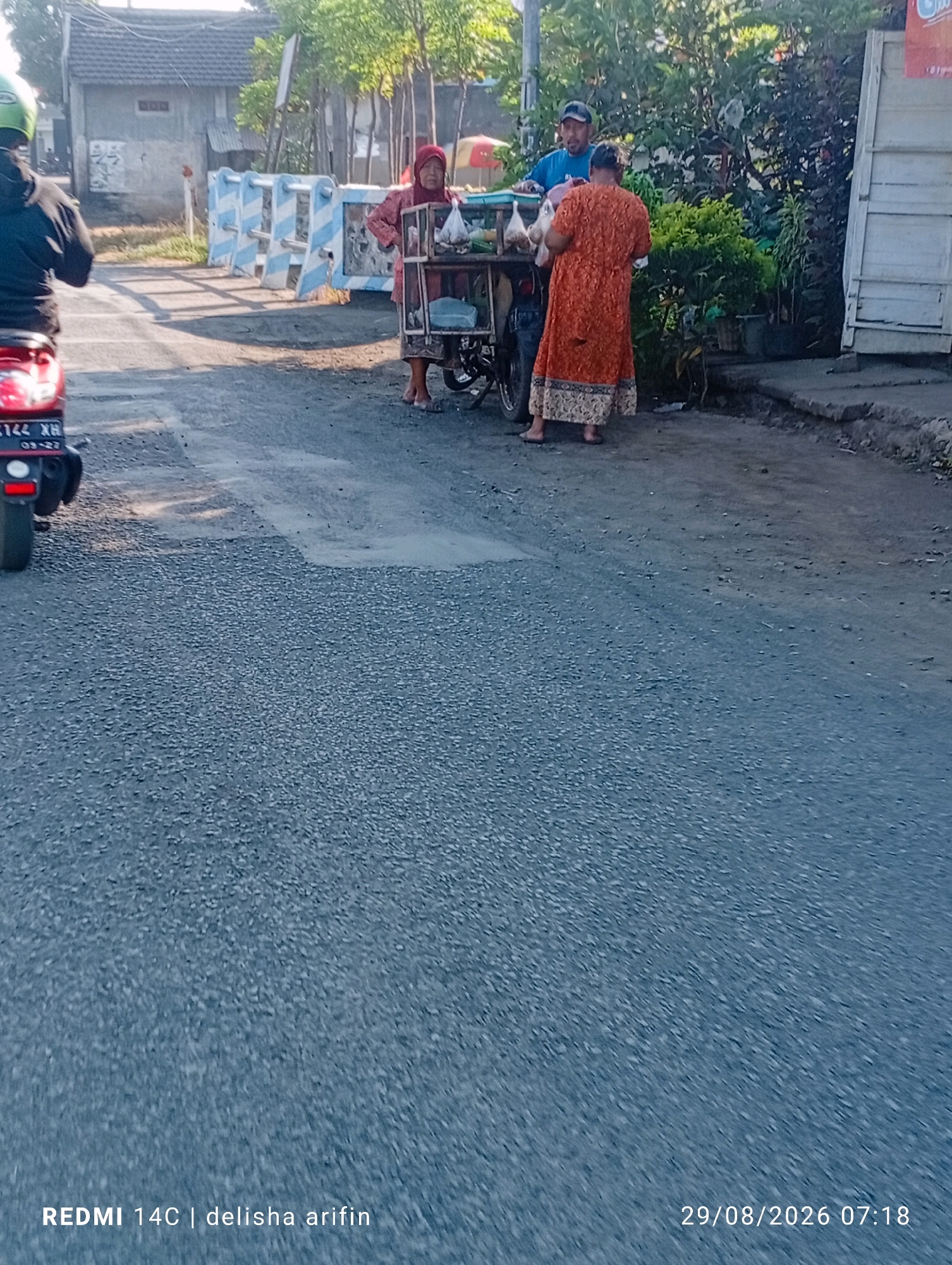 In Plinggisan, Indonesia, a street scene unfolds with a motorcycle parked on the left side of the image. A vendor with a cart is engaged with two customers. The vendor, wearing a pink headscarf and top, stands behind the cart, appearing to be interacting with a person on the right. This person, dressed in an orange dress, faces away from the camera, and is handing something to the vendor or receiving something from the cart. A third individual, a man in a blue shirt and cap, stands behind the vendor, also near the cart. The environment is paved with asphalt, with foliage and a building visible in the background. The image is dated 29/08/2026 at 07:18.