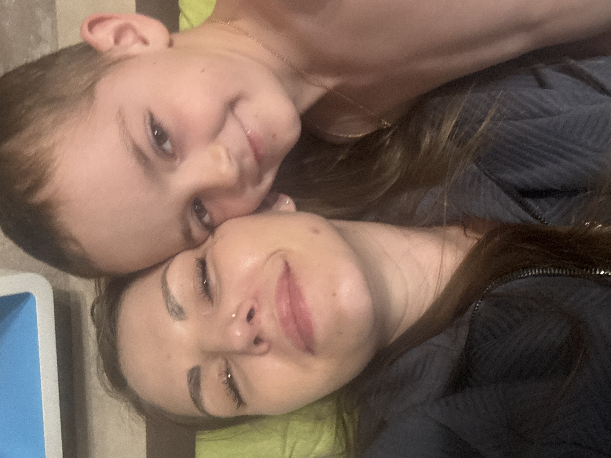 A close-up shot depicts two people lying down next to each other. The individual on the bottom appears to be a woman with brown hair and a nose piercing. Her eyes are closed and she is slightly smiling. The second individual, who appears to be a child, is positioned above the woman. The child is looking at the camera with a slight smile. The location is unknown and there is no evidence to suggest it is in the city of Раздолье, Russia.