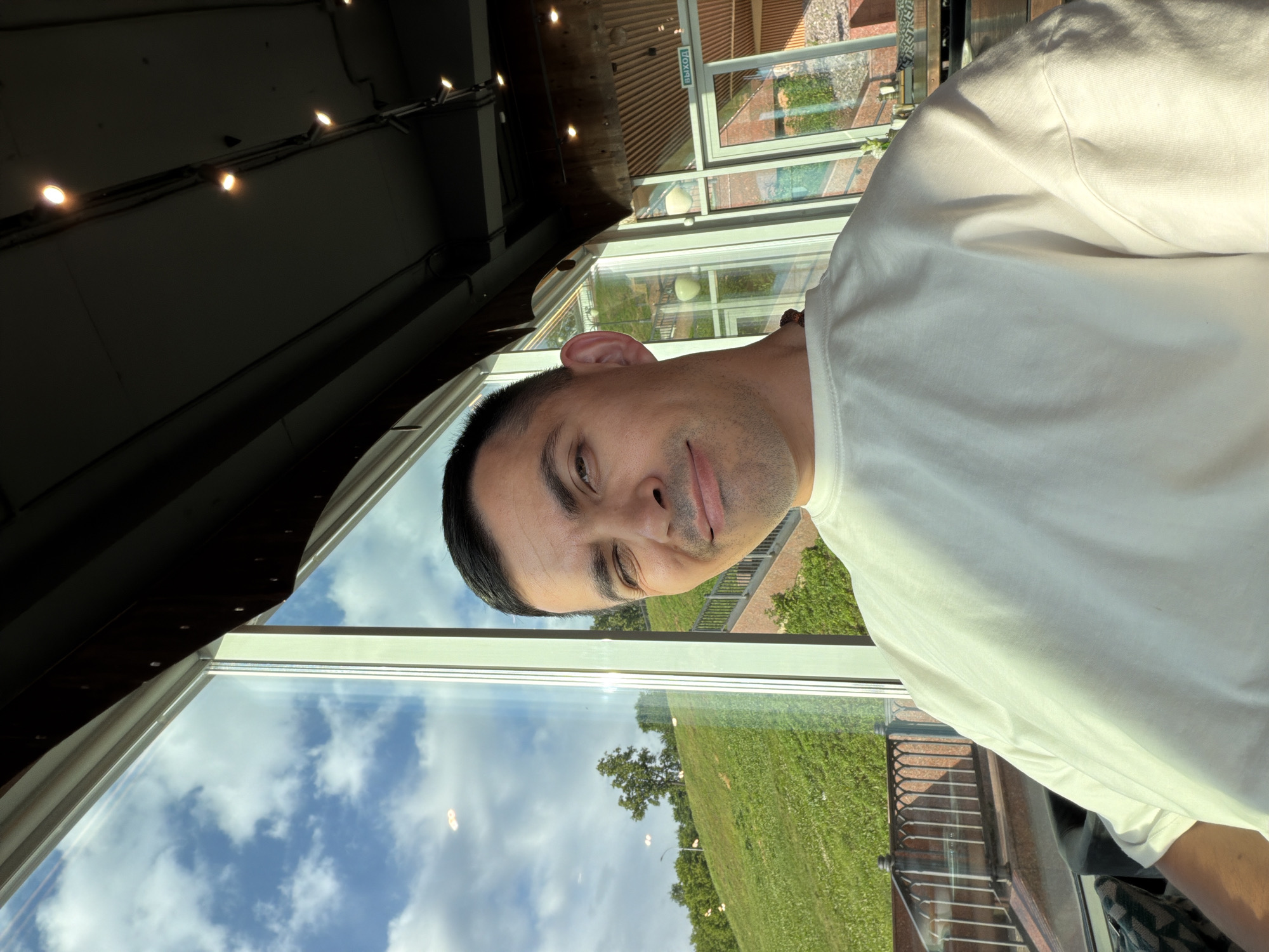An overhead shot captures a man from the chest up, selfie-style. He is looking slightly to his right and winking with his left eye. He is wearing a plain white t-shirt and a dark necklace. The man is positioned in front of a large window that overlooks a green, grassy hill with trees and a cloudy sky. The window frame is light-colored. In the background, through the window, there are glimpses of a building with wooden interior design and a sign with Russian text that says "ВЫХОД" which translates to "EXIT." The lighting suggests it is daytime with sunlight streaming through the window, creating reflections on the glass and illuminating the man's face and shirt. The ceiling above is dark with visible lights. The location appears to be a modern establishment with large windows, possibly a cafe or restaurant, overlooking an outdoor green space in Izhevsk, Russia.