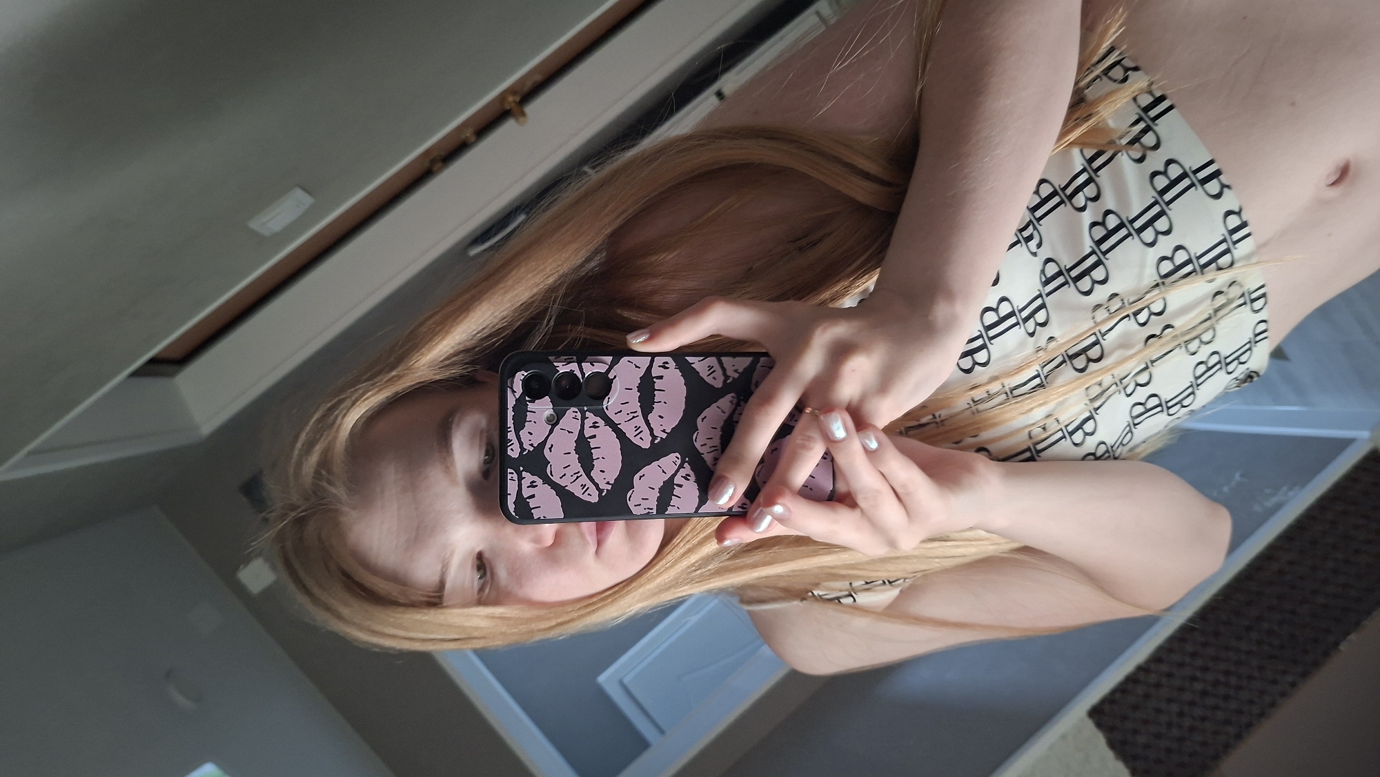 A young woman with long, light blonde hair is taking a selfie in a mirror. She is wearing a light-colored top with a black pattern of repeating "B" logos. The phone case is black with a pink lip print design. The background appears to be a room with a ceiling and a cabinet. The location is in the city of Chaikovsky, Russia. No further details can be inferred from the image.