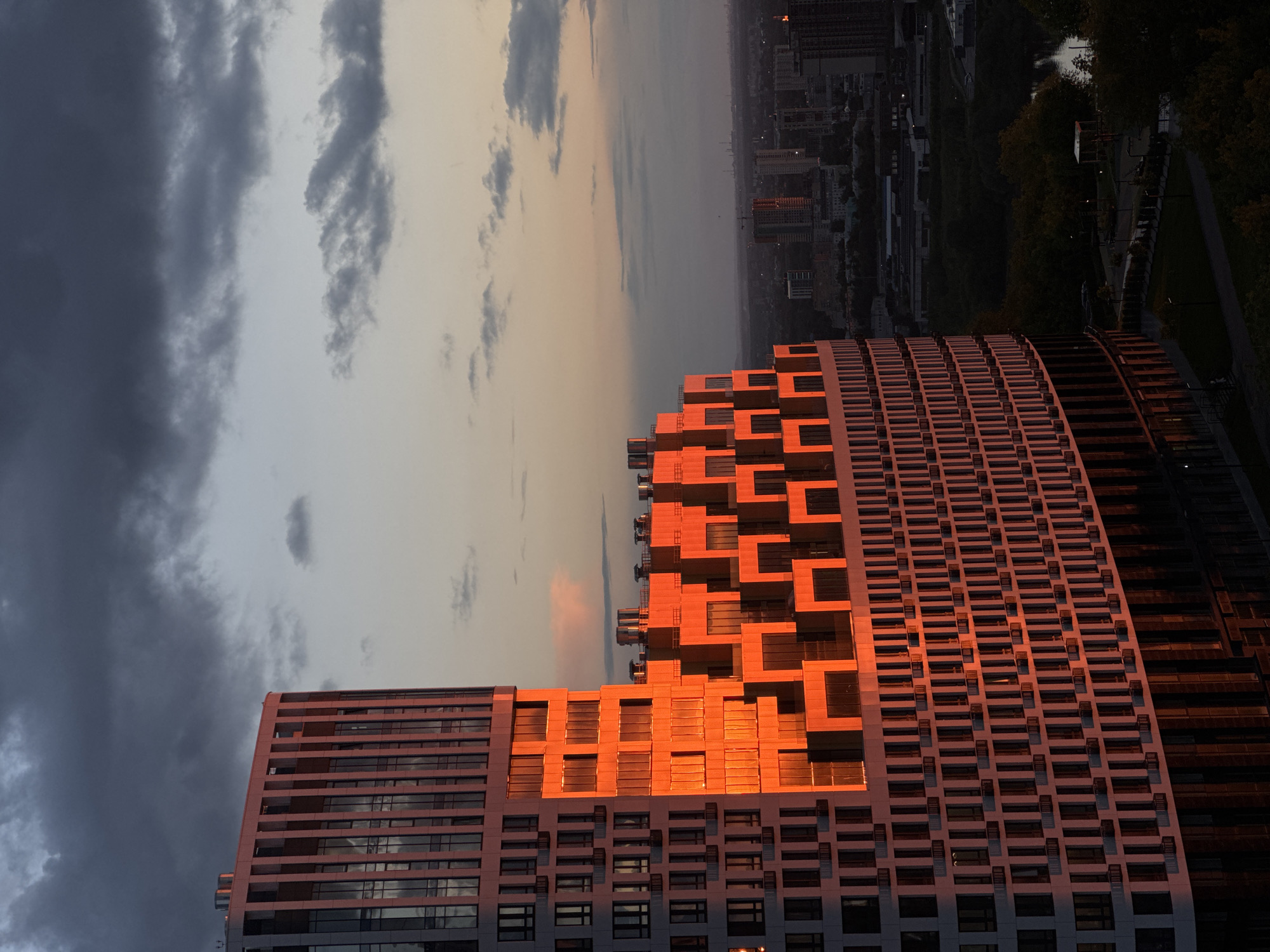 An aerial shot looking across a city towards the sunset. The large building on the lower half of the shot is a modern design building with a rectangular base and angled/staggered design on top. The sunlight is reflecting on the building making it appear orange. The city is to the top half of the shot, with green trees and a body of water to the top right. The sky has some storm clouds on the left, and an orange hue closer to the city.