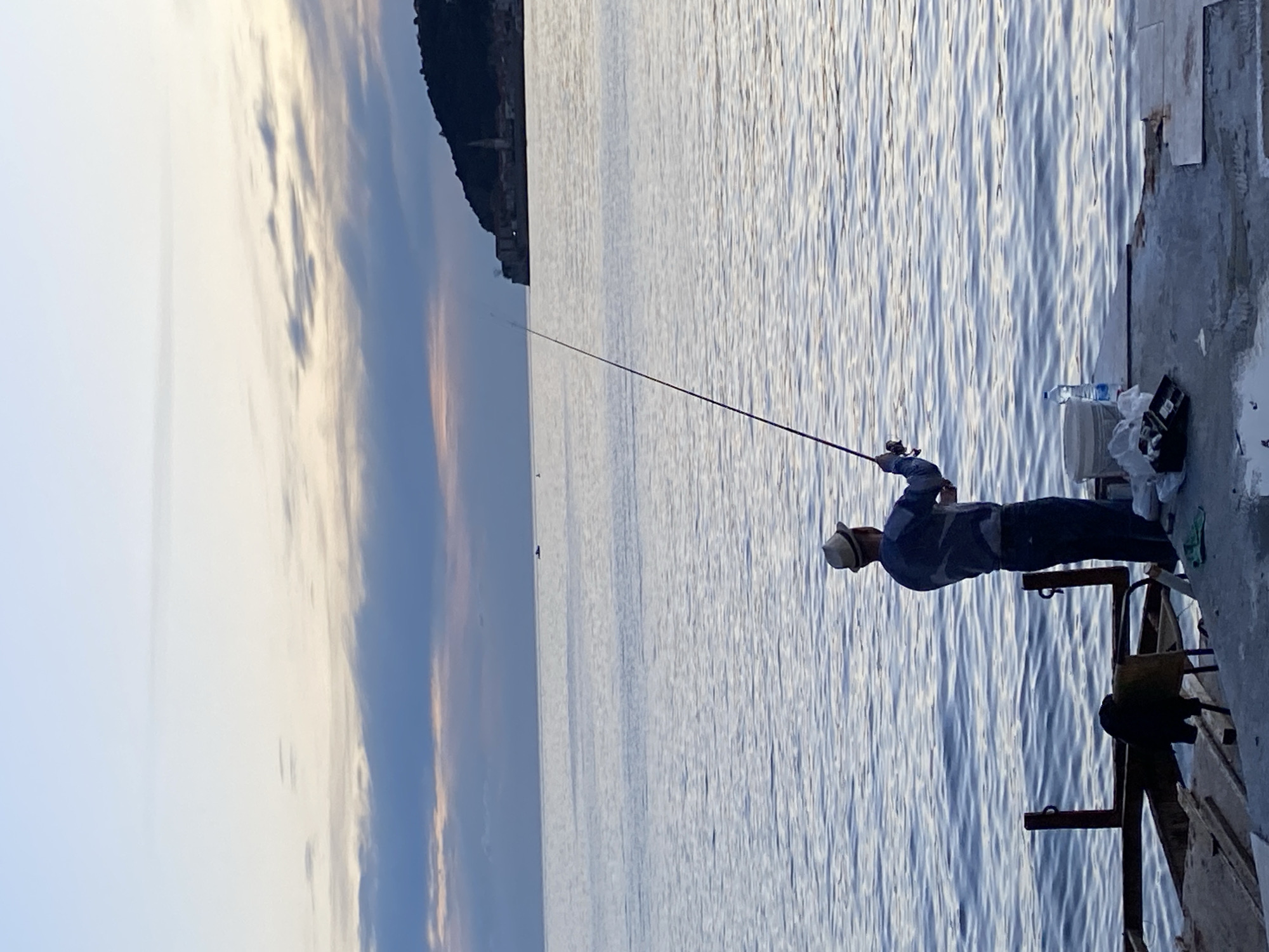 A man is fishing off a concrete pier in the city of Budva, Montenegro. He stands near the edge of the pier, holding a fishing rod that extends over the water. Beside him are various items, including a water bottle, a bucket, and a bag. The water is rippled and reflects the sky, which has varying shades of blue and hints of orange near the horizon. In the background, there is a strip of land with buildings visible along the coast. The overall scene suggests a calm, outdoor setting, focused on the activity of fishing.