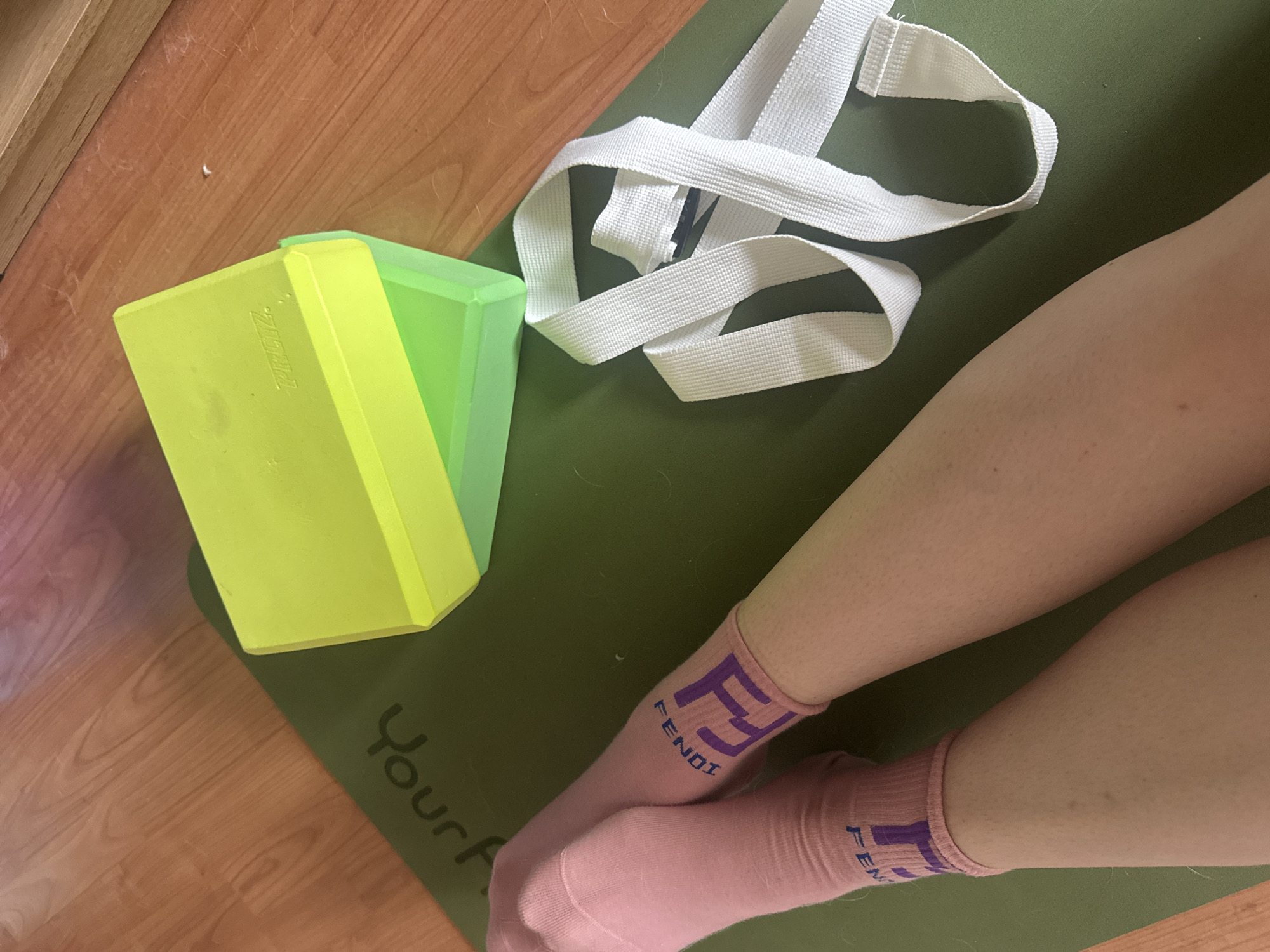 An overhead, medium shot captures a pair of pink socks with purple "FENDI" logos on a green yoga mat. The socks are worn by a person whose legs are visible. A white yoga strap is coiled to the right of the socks. Two lime green yoga blocks are stacked on the upper right corner of the yoga mat, which is positioned on a wooden floor. The background consists of the wooden floor and a piece of furniture.