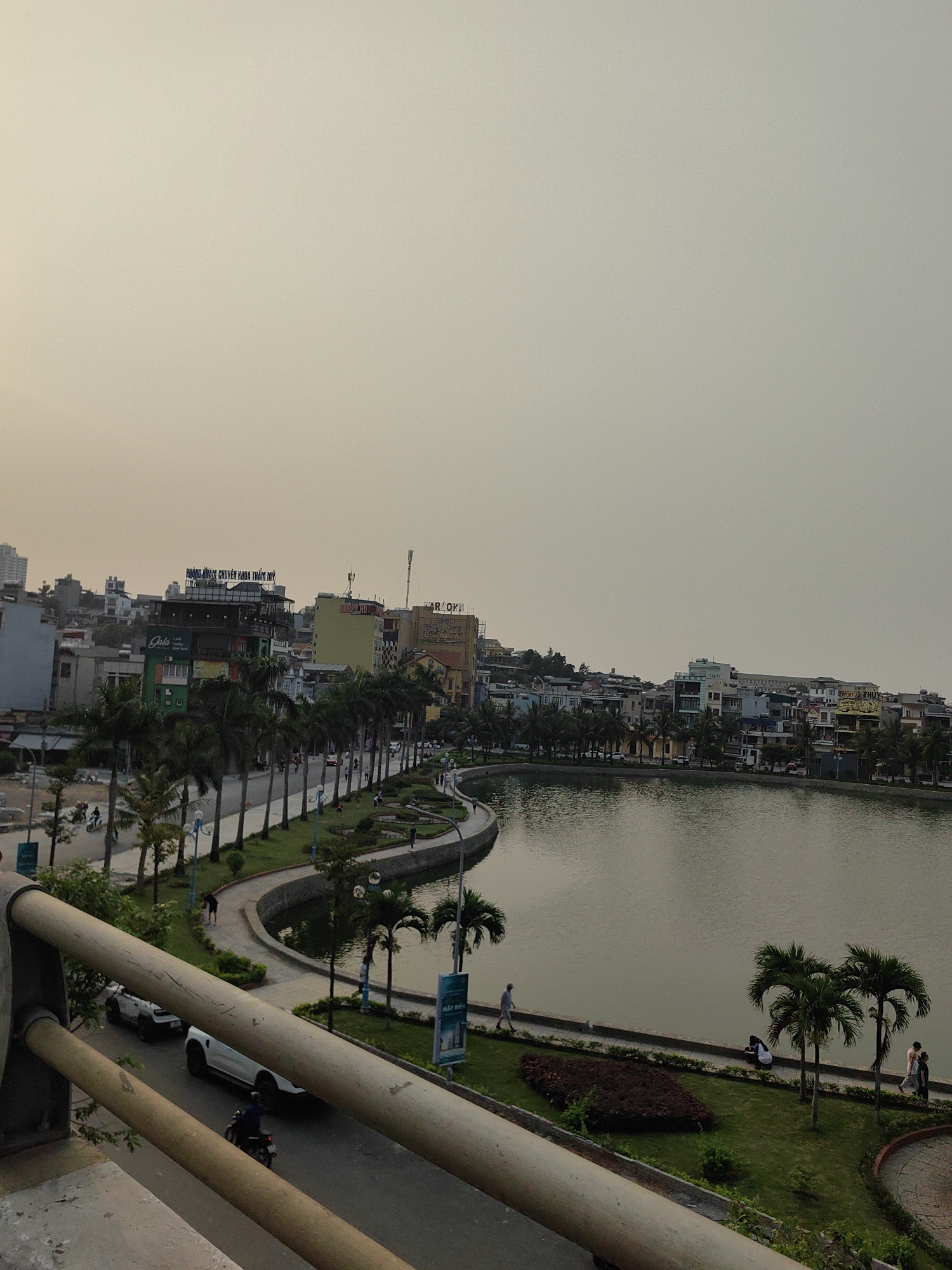 The image shows a daytime view of Hong Gai Ward in Vietnam. A large body of water with a winding edge occupies the right half of the frame, lined with palm trees. A pedestrian walkway runs along the water's edge. Buildings are visible in the background, indicating urban development. A roadway is present in the lower portion of the image, with a few cars and a motorbike. A bridge railing is visible in the foreground, suggesting the photo was taken from a bridge. Several people can be seen walking, sitting, or standing along the water's edge. The sky appears overcast.