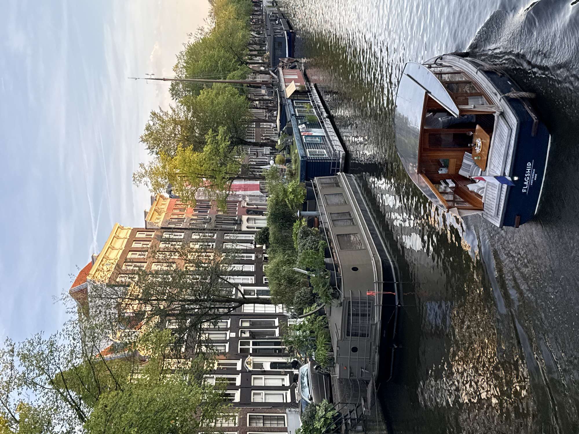 This image depicts a canal in Amsterdam, Netherlands, likely during the late afternoon or early evening, as indicated by the golden light illuminating the buildings. In the foreground, a dark blue motorboat named "FLAGSHIP" with "AMSTERDAM" written on its stern is moving away from the viewer, leaving a wake in the water. The boat has a wooden interior, a small table with bottles and glasses, and seating cushions. To the left of the motorboat, a large, long houseboat with a green roof and numerous potted plants is docked. Beyond this houseboat, a row of traditional Amsterdam canal houses with red-tiled roofs and dark facades are visible. Further down the canal, other houseboats are moored, and trees line the canal banks. The sky is a pale blue with wispy clouds and contrails.