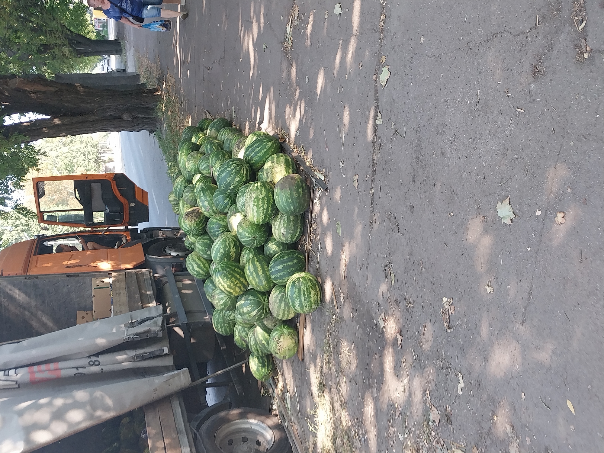 The image shows a pile of watermelons on the side of a street in Dnipro, Ukraine. A large truck with an open door is parked next to the watermelons. A woman is walking down the sidewalk with shopping bags. The scene appears to be a street market or a roadside stand.