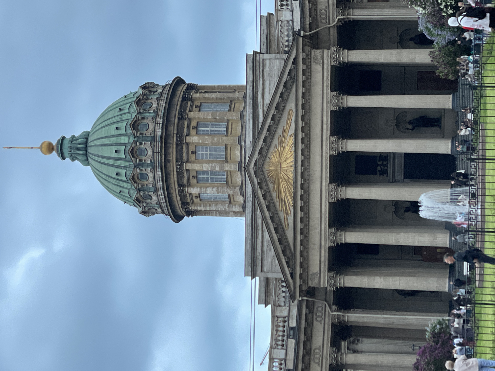 This image shows the Kazan Cathedral in Saint Petersburg, Russia. The building features a large green dome topped with a golden spire. Adjacent to the dome is the main facade of the cathedral, with a triangular pediment adorned with a golden sunburst. Below the pediment are numerous columns forming a colonnade, typical of neoclassical architecture. The foreground includes a fence and a view of people, vegetation, and a fountain. The sky is overcast.