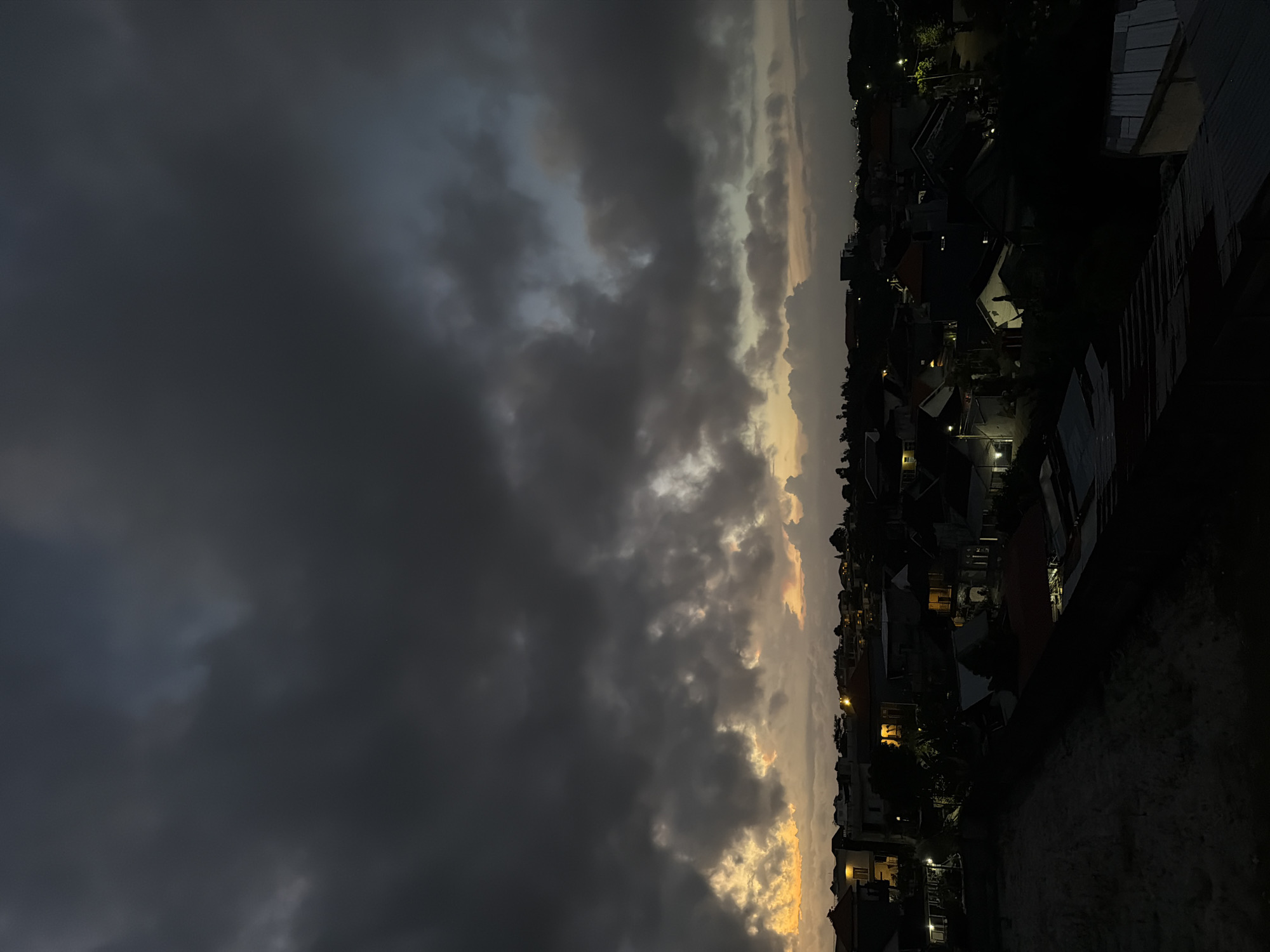 The image depicts a cityscape at twilight, likely in Kuta Selatan, Indonesia. The foreground contains the dark, silhouetted rooftops of residential buildings with occasional light emanating from windows and streetlights. In the distance, a dramatic sky is filled with dark, ominous storm clouds, tinged with the golden hues of the setting sun. The photograph is oriented vertically, giving a sense of height and scope. No people are visible. The overall impression is a blend of urban development juxtaposed against a backdrop of natural atmospheric phenomena.