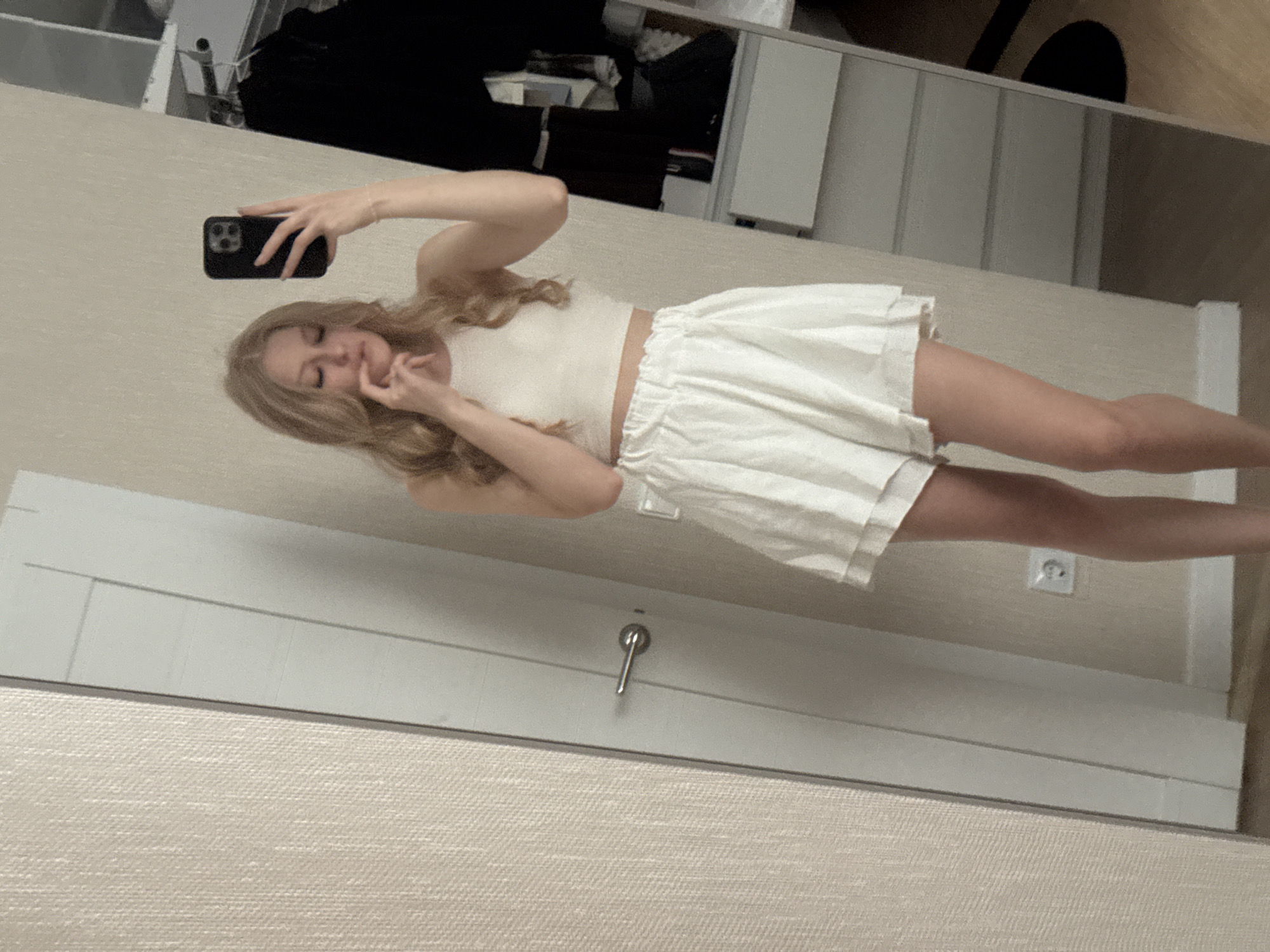 A young Caucasian woman with blonde hair stands in a room, taking a mirror selfie. She is wearing a white crop top and white shorts. Her left hand is raised to her face, and she holds a black smartphone in her right hand. The background shows a door on the left, a wall with textured wallpaper, and a closet with clothes on the right. The scene appears to be indoors, possibly a bedroom or dressing area. No specific elements suggest a location in the city of Perm, Russia.