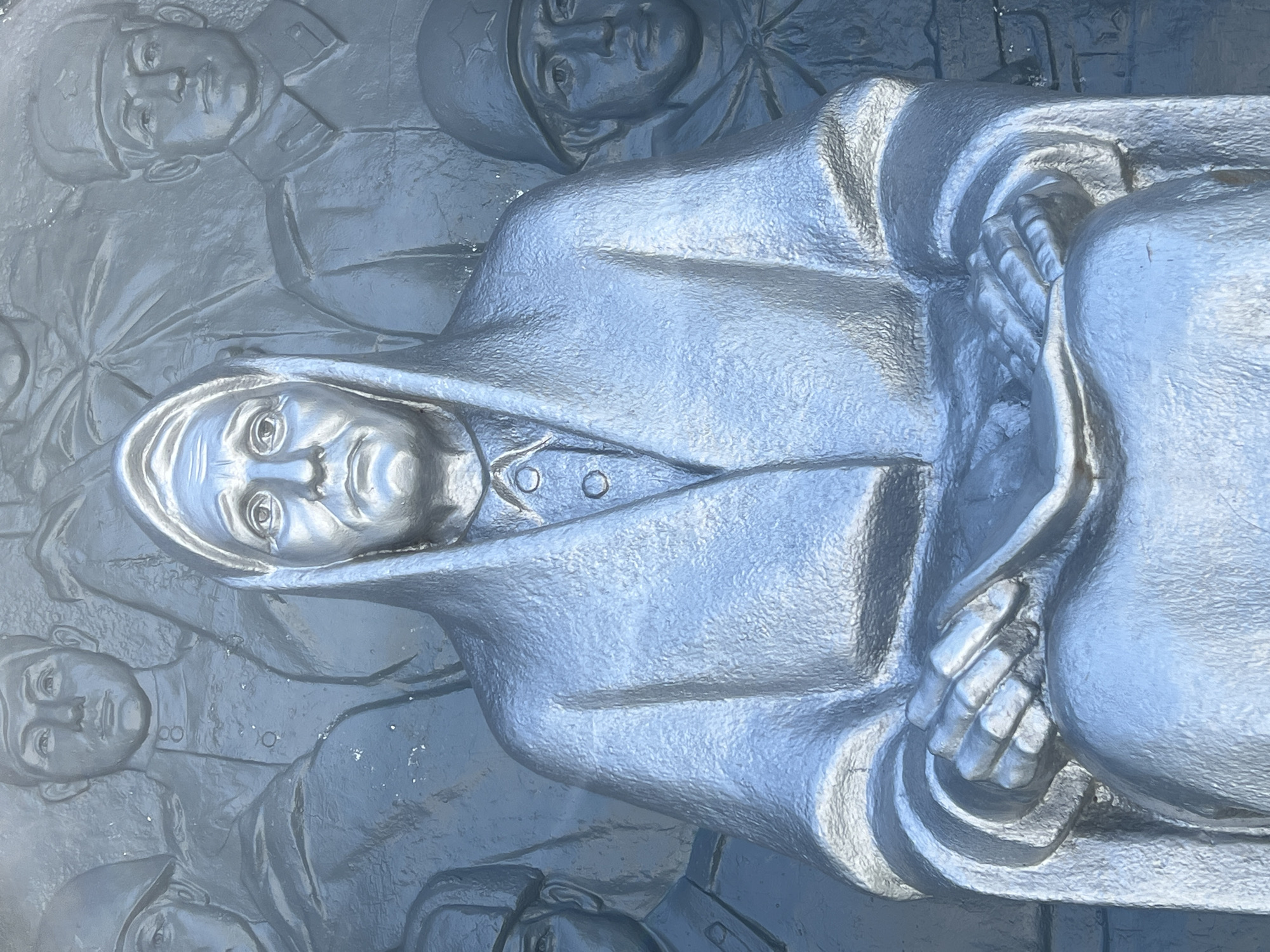 The image depicts a close-up of a silver-colored relief sculpture. The central figure is an elderly woman wearing a headscarf and a coat. Her expression appears somber. Her hands are clasped in her lap, holding what seems to be a piece of fabric. Flanking her on either side are reliefs of soldiers in uniform, some wearing helmets. The background is a textured surface, also rendered in silver. The sculpture is likely part of a larger monument, possibly commemorating historical events or individuals. The location is identified as the city of Takhtamukai, Russia, suggesting the sculpture may be related to the region's history.