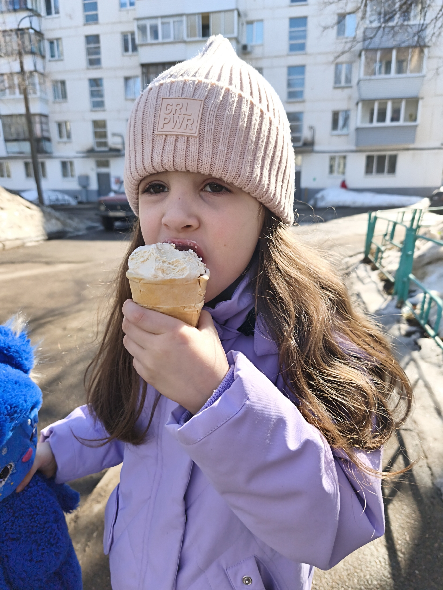 A young girl is centrally positioned outdoors in a residential area of Chaikovsky, Russia, actively consuming a scoop of vanilla ice cream from a waffle cone. She wears a light pink ribbed knit beanie with a "GRL PWR" patch on the forehead and a light purple quilted jacket. Her long, dark brown hair frames her face. Her right hand holds the ice cream cone, which she is licking or biting with an open mouth. A blue fuzzy plush toy with prominent eyes is partially visible in her left hand on the lower left. The background features a multi-story, white apartment building with numerous windows and balconies, consistent with post-Soviet residential architecture. The ground is a dirt or asphalt path with visible patches of melting snow and ice, suggesting a late winter or early spring season. A green metal fence is present on the right, and a dark-colored vehicle is partially visible in the mid-ground. The scene is brightly lit by direct sunlight.