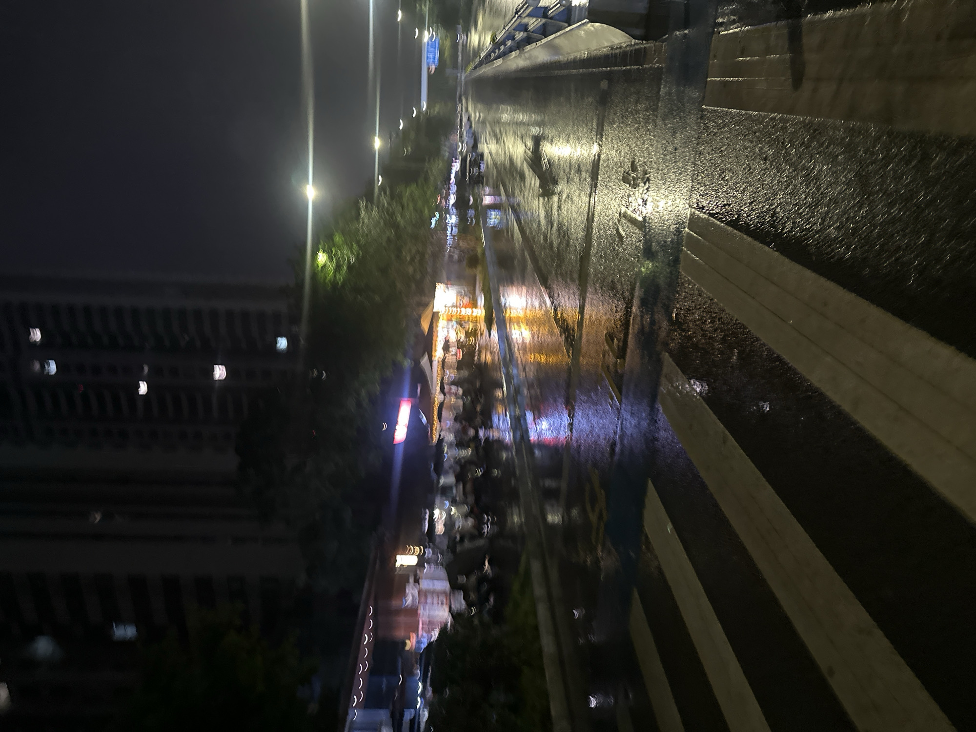 The image depicts a nocturnal urban scene in Chongqing High-tech Zone, China, captured from a low vantage point looking down a wet street. The foreground features a wide pedestrian crosswalk with distinct white and dark stripes, reflecting overhead light sources. The asphalt road surface is thoroughly wet, creating numerous bright, elongated reflections of streetlights and various colorful lights from establishments.
To the left of the road, a series of small commercial establishments, possibly shops or food stalls, are brightly illuminated with yellow, orange, red, and purple lights, which produce prominent, blurred reflections on the wet ground. Above these establishments, blurred green foliage from trees or bushes is visible. Beyond the establishments, tall, dark multi-story buildings with scattered illuminated windows rise into the dark sky.
The road extends into the midground and background, flanked on the right by a low blue and grey barrier or railing. Several overhead streetlights are visible, producing bright, streaky flares due to the low-light conditions and motion blur. In the far background, a large, lighter-colored structure, possibly an overpass or bridge, spans horizontally across the upper part of the road. The overall atmosphere is characterized by artificial light reflecting off wet surfaces in an urban environment at night. No distinct individuals or specific actions are clearly discernible due to blur and distance.