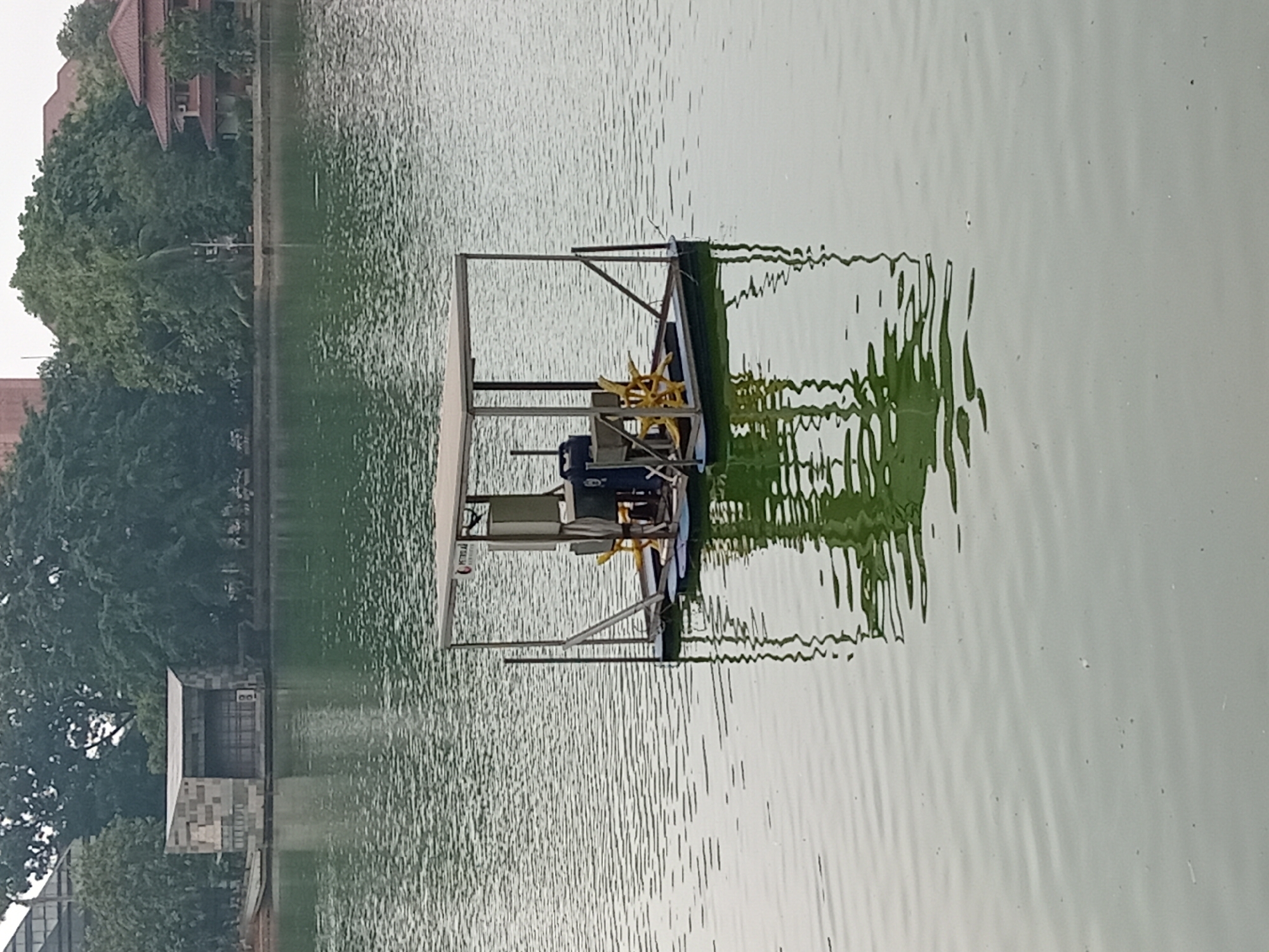 In Depok, Indonesia, a water aerator sits in the middle of a calm, green body of water, likely a lake or large pond. The aerator, a floating platform with a metal frame and a canopy, houses machinery and paddle wheels designed to oxygenate the water. The water's surface is slightly rippled, reflecting the sky and the distant shoreline. The background features lush green trees and architectural structures, including a modern-looking building with a gray facade on the left and a building with a red tiled roof on the right, suggesting an urban or park setting. The overall atmosphere is serene and still.