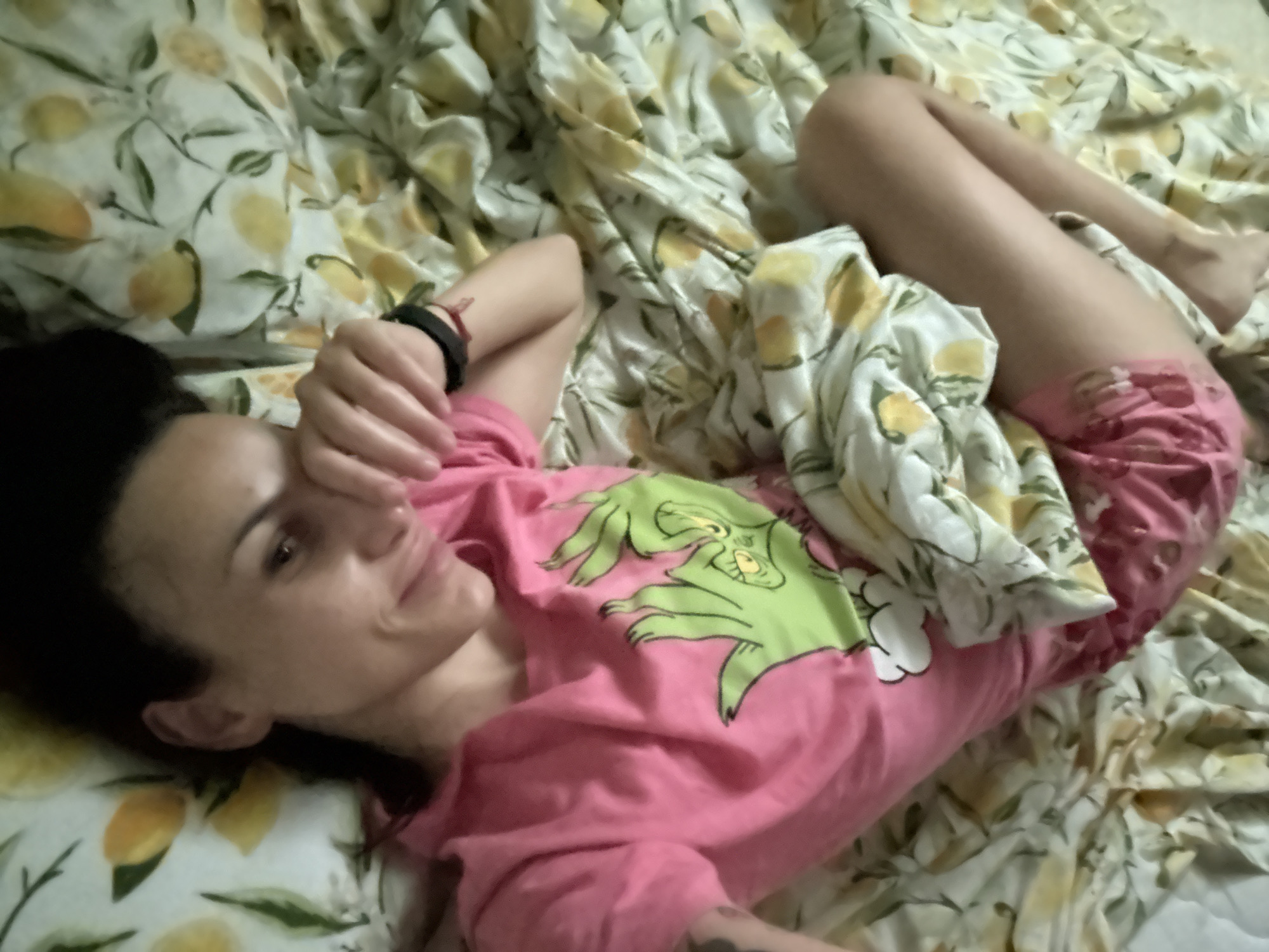 A woman is lying on a bed with a lemon-patterned sheet and pillowcases. She is wearing a pink shirt with a Grinch design and pink shorts. Her right arm is raised, and her hand is partially obscuring her face. Her legs are bent at the knees. The location is most likely inside a residence; there is no clear indication of it being in the city of Pleven, Bulgaria.