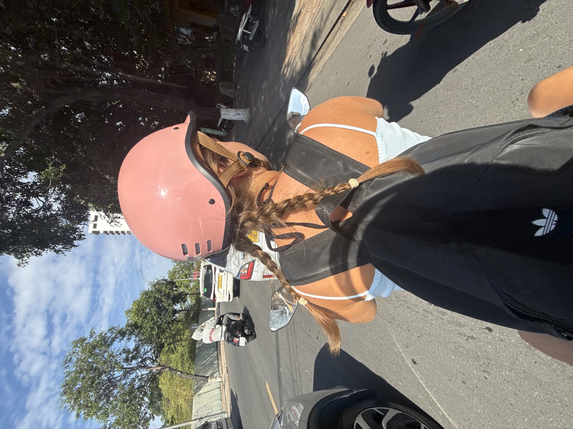 This is a first-person perspective photo taken from the back of a motorcycle in Nha Trang, Vietnam. The rider is wearing a pink helmet and a light blue tank top, with a black backpack. Their hair is in two braids. The motorcycle is on a paved road with traffic, including other motorcycles and cars. The scene is outdoors during the day, with clear blue skies and sunlight casting shadows. The background shows trees, buildings, and a white fence. The image captures a typical street scene in a Vietnamese city.