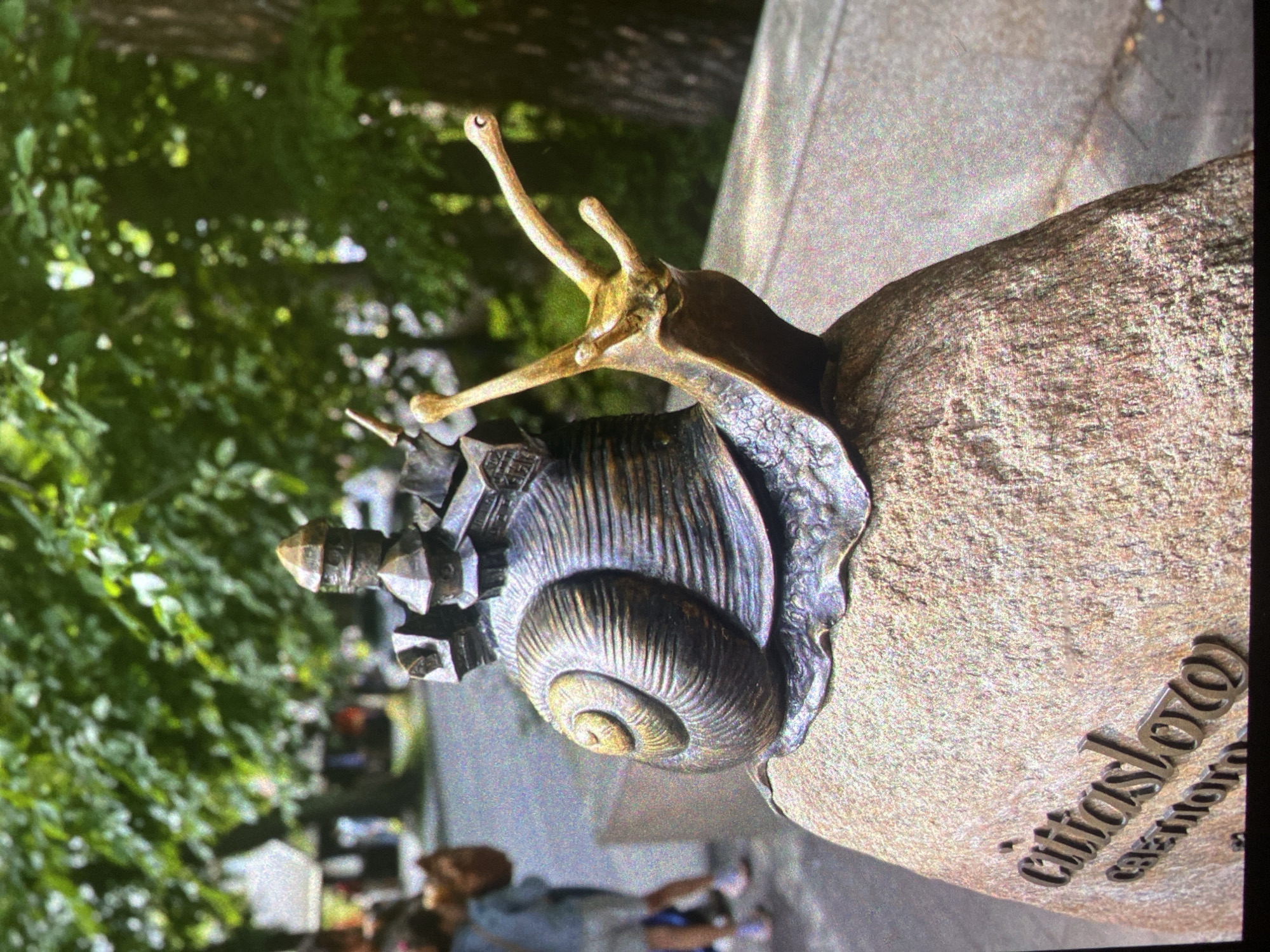 A close-up shot depicts a bronze sculpture of a snail. On top of the snail's shell is a detailed replica of the Admiralty building in St. Petersburg. The sculpture rests on a granite pedestal inscribed with Russian text. In the background, out-of-focus, are trees and pedestrians, suggesting the sculpture is in a park or public space. The image is taken from a slightly low angle, emphasizing the size and detail of the sculpture. The location is likely in Saint Petersburg, Russia, based on the Admiralty building replica.
