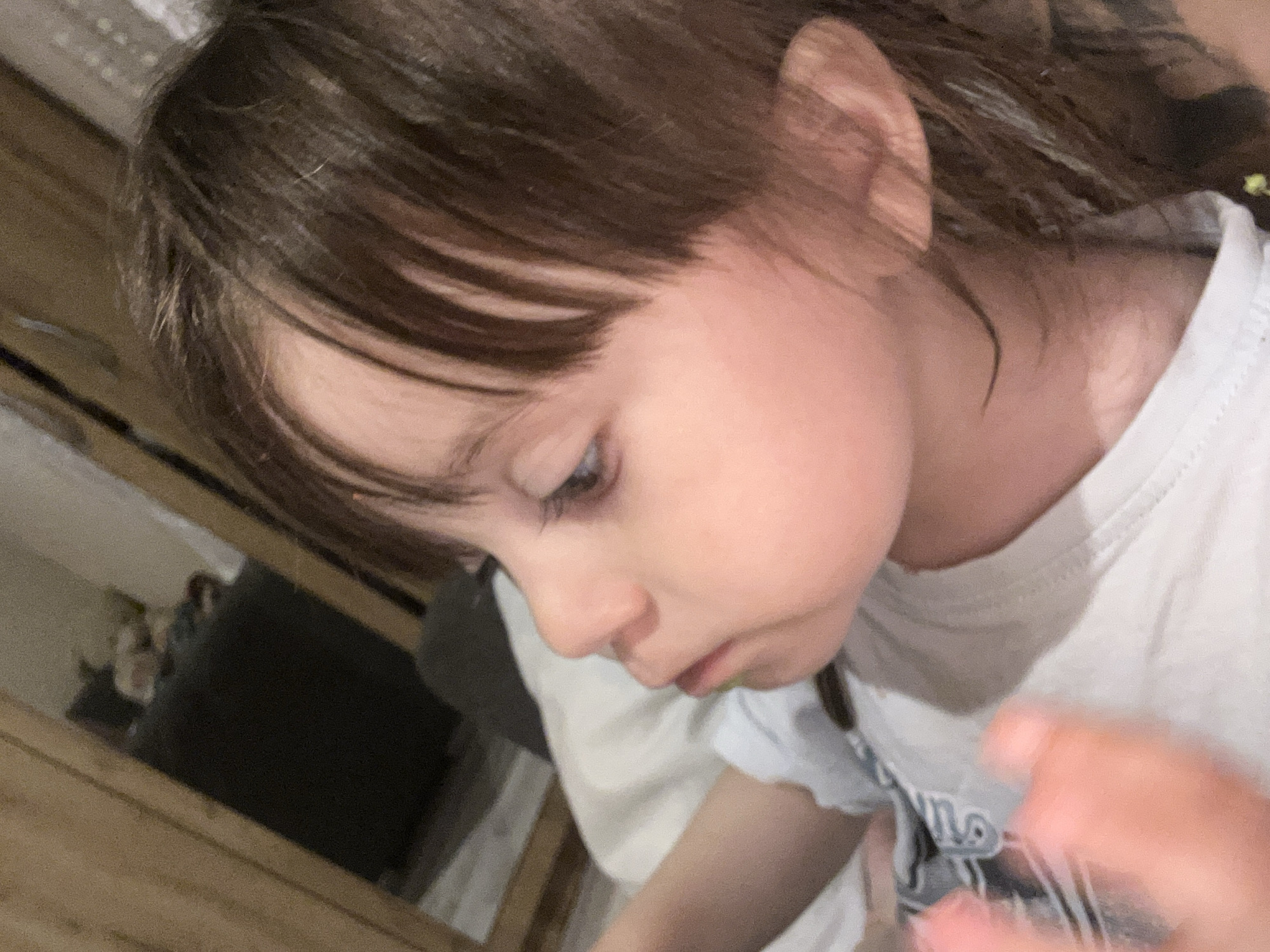 A close-up, slightly angled shot features a young child. The child has fair skin, brown hair with bangs partially covering the forehead, and is wearing a white T-shirt. The child's gaze is directed downwards, suggesting focus on something out of frame.
The background shows portions of what appears to be a room, including sections of a wooden surface or furniture on the left side. A blurry object and hand are visible on the bottom right. There is no information in the image to suggest that the photo was taken in the city of Tsarevo, Bulgaria.