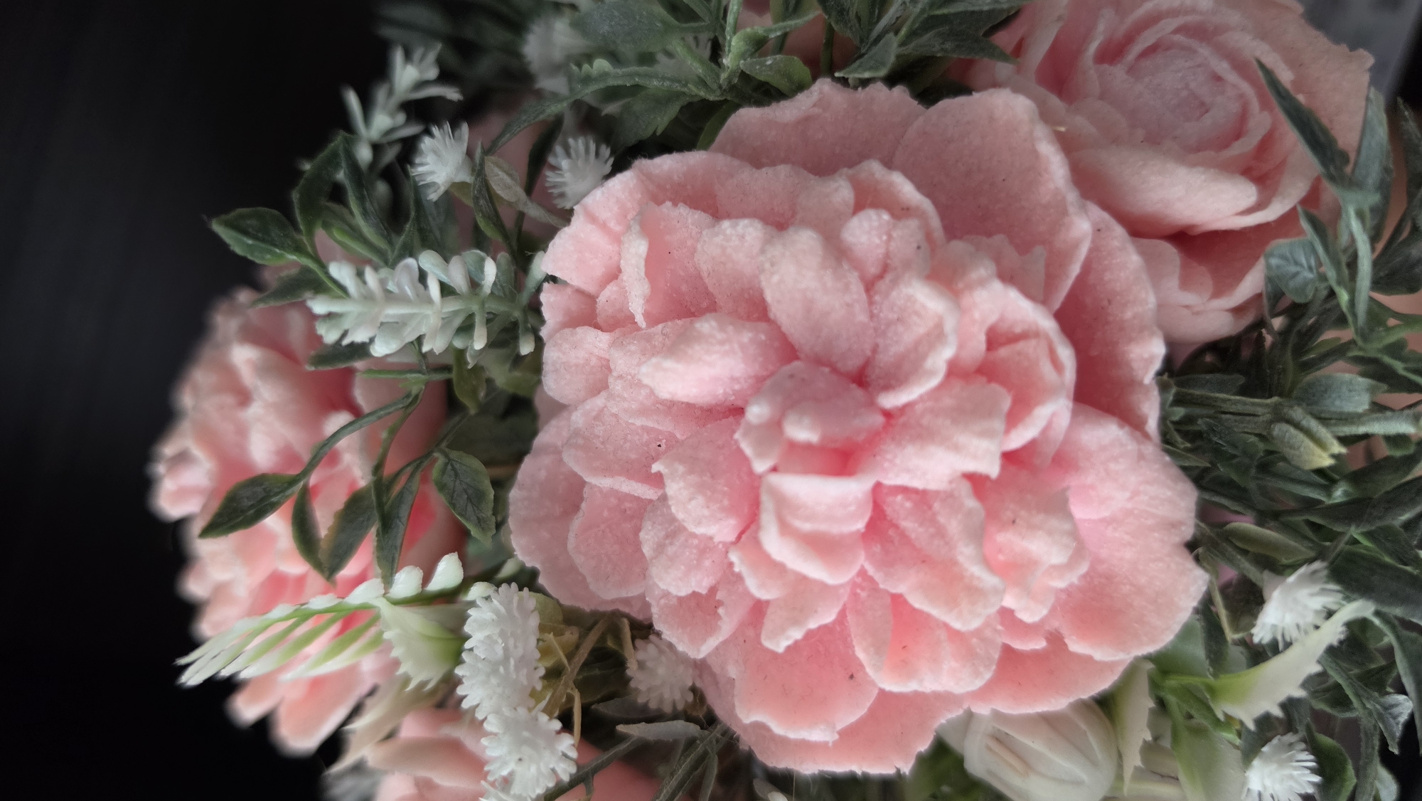 A close-up, detailed, macro photograph captures a pink, velvety flower, possibly a peony, in the foreground. The flower is densely packed with numerous ruffled petals, each with a slightly fuzzy texture and a light dusting of what appears to be glitter or sugar. Surrounding the central flower are smaller white floral accents and lush green artificial foliage, which frame the pink bloom. The background is a dark, blurred void, creating a strong contrast that emphasizes the intricate details of the flower. The image is professionally lit, highlighting the texture and vibrant color of the subject. There is no indication of any specific location within Jūrmala, Latvia.