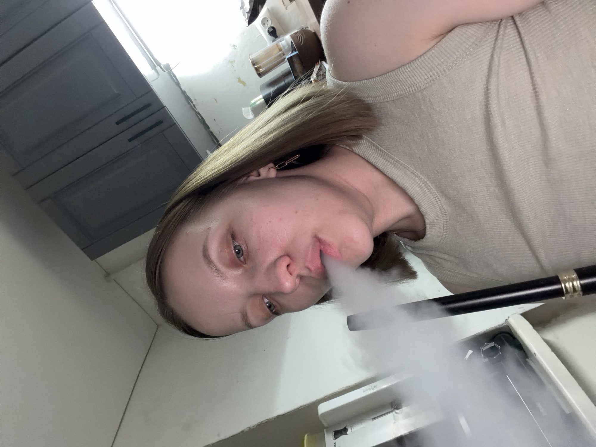 A person, likely female, is exhaling white vapor, possibly from a vaping device or hookah. She has light skin, blonde hair, and is wearing a light-colored top. The background includes kitchen cabinets and various household items. The specific location in Reshetnikovo, Russia is not discernible from the image.