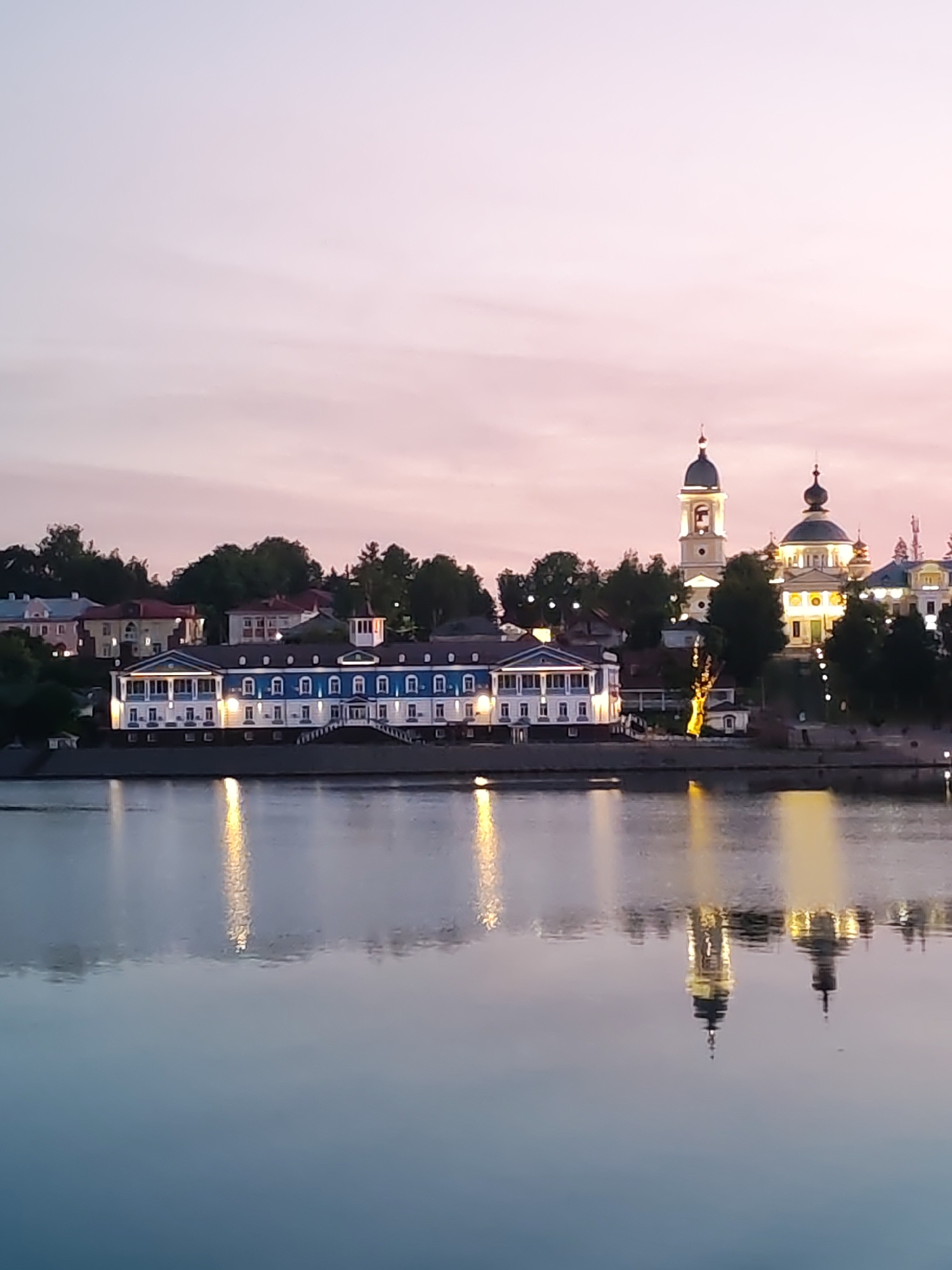 The image displays a panoramic view of a city waterfront at either dawn or dusk. The sky above transitions from a light blue at the zenith to a soft pink and purple gradient towards the horizon. In the foreground, a calm, dark body of water, likely a river, occupies the lower third of the frame, reflecting the ambient light and illuminated structures.
Along the far bank of the river, a series of buildings are situated on a slightly elevated terrain. To the left, a prominent multi-story building features a dark blue pitched roof, white facades, and numerous illuminated windows. A grand staircase with multiple landings descends from this building towards the water's edge. Trees and other residential-style buildings with varied architectural styles are visible further up the bank behind this central structure.
Towards the right side of the image, two distinctive structures dominate the skyline. A tall, light-colored bell tower with a dark, onion-shaped dome and finial is brightly illuminated. Adjacent to it stands a larger, ornate building with multiple domes, also brightly lit, suggesting a religious edifice such as a church or cathedral. These structures are nestled among dark green trees.
Artificial lights from the buildings cast elongated, shimmering reflections of yellow and white onto the tranquil water surface, including clear inverted images of the bell tower and domed building. No individuals are discernible within the frame. The scene captures an urban or semi-urban environment during twilight, characterized by architectural illumination and still water reflections. The specific city and location within "Unknown, Unknown" are not identifiable from the image.