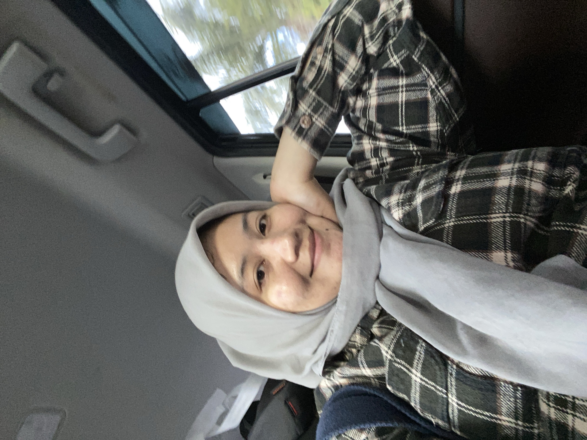 A medium shot captures a woman wearing a light gray hijab and a black and white plaid flannel shirt, resting her head on her right hand. She is seated inside a vehicle, evidenced by the car's interior visible in the background, including a handle on the ceiling and a window with a blurred outdoor scene of trees and sky. The woman's gaze is directed towards the camera, and she has a gentle smile. The environment suggests a journey, likely within the city of Banjarbaru, Indonesia, given the naturalistic outdoor scenery visible through the window.