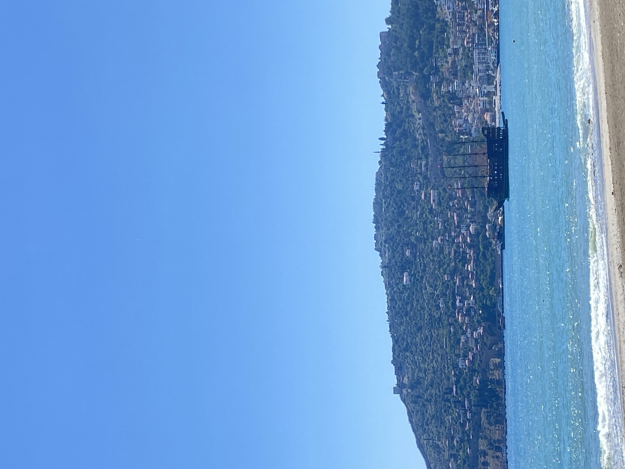 A daytime photograph shows Alanya, Turkey, from a beach perspective. A mountain range densely populated with buildings and trees occupies the upper right portion of the frame, adjacent to the beach. The shoreline meets a blue body of water with mild waves breaking onto the sand. A pier-like structure extends out over the water. The left portion of the image consists of a clear, bright blue sky.