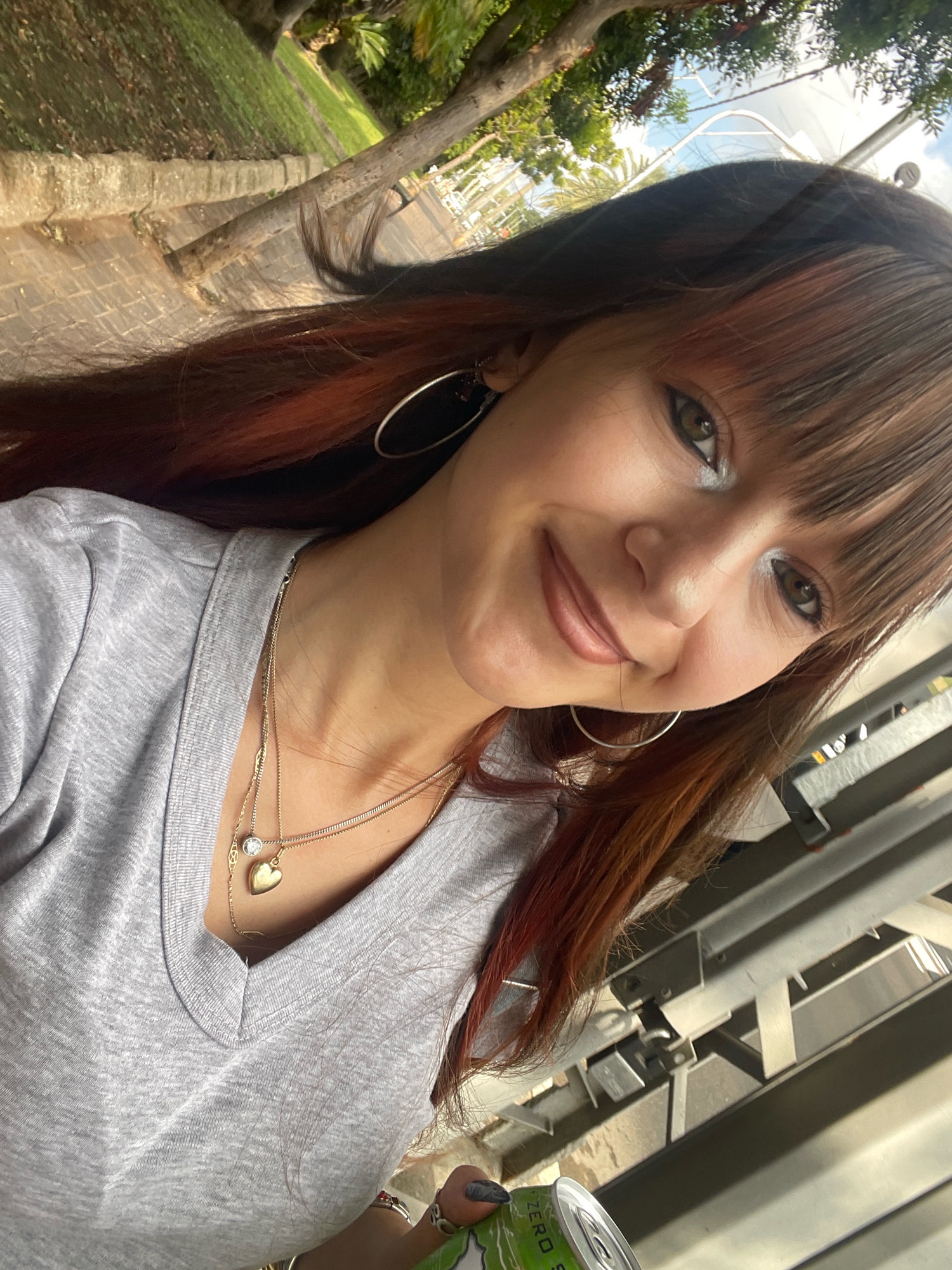 A young woman with long, auburn-streaked dark hair and bangs is taking a selfie outdoors. She is wearing a grey V-neck t-shirt and two necklaces, one with a clear pendant and another with a gold heart pendant. She is also wearing hoop earrings. Her makeup includes dark eyeliner and eyeshadow. She is holding a green can in her right hand.
The background suggests an urban or park-like setting with trees, a paved walkway, and some metal structures. The lighting indicates it is daytime, with sunlight casting shadows. The angle of the shot implies she is standing or possibly leaning. The image appears to be taken in Raanana, Israel, given the context provided.