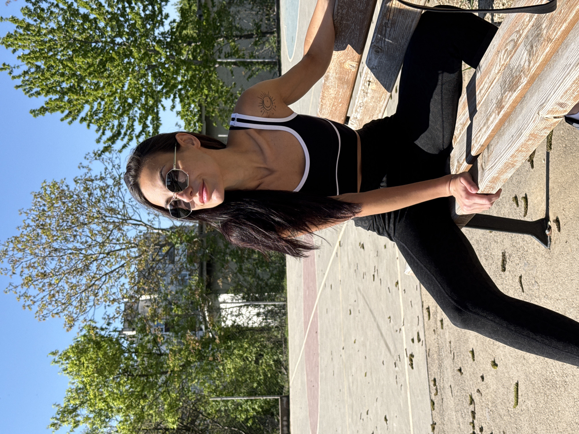 A woman in a black athletic top and black leggings sits on a wooden bench outdoors. The image appears to have been taken in Pleven, Bulgaria, given the surrounding cityscape and foliage. The woman is wearing sunglasses. A tattoo is visible on her upper arm. She is holding the bench with her hands. The ground is concrete with scattered debris. Trees and buildings are visible in the background under a blue sky. The lighting suggests it is daytime.