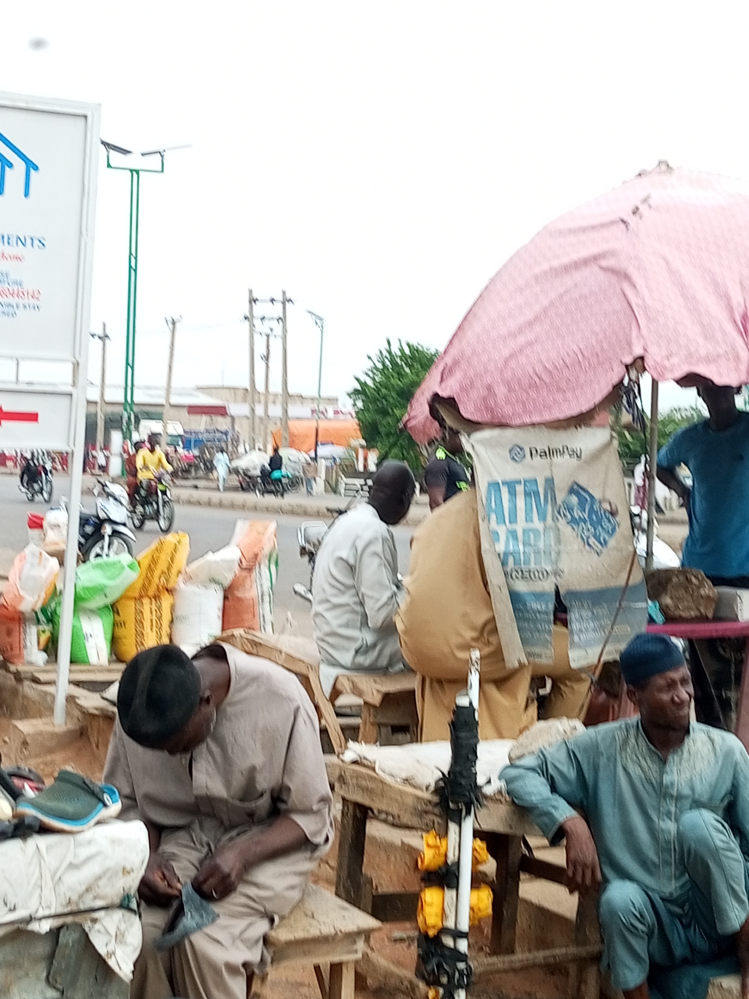 The image depicts a street scene in Gombe, Nigeria, likely a marketplace or busy roadside. In the foreground, three men are present. One man, wearing a dark cap and a light brown traditional outfit, is seated on a wooden bench and appears to be repairing a shoe. Another man, wearing a blue cap and a light blue traditional outfit, is also seated, looking off to the right. A third man, in a tan traditional outfit, is seated behind a large, light-colored bag with "PalmPay ATM CARD" printed on it, partially covered by a pink umbrella. In the background, the street is visible with traffic, including motorcycles and a few people walking. Several stalls or vendors are set up along the roadside, displaying various goods, including sacks of what appear to be grains or cement, and some plastic containers. The environment suggests a typical urban setting in Nigeria, with a mix of commercial activity and public infrastructure like utility poles.