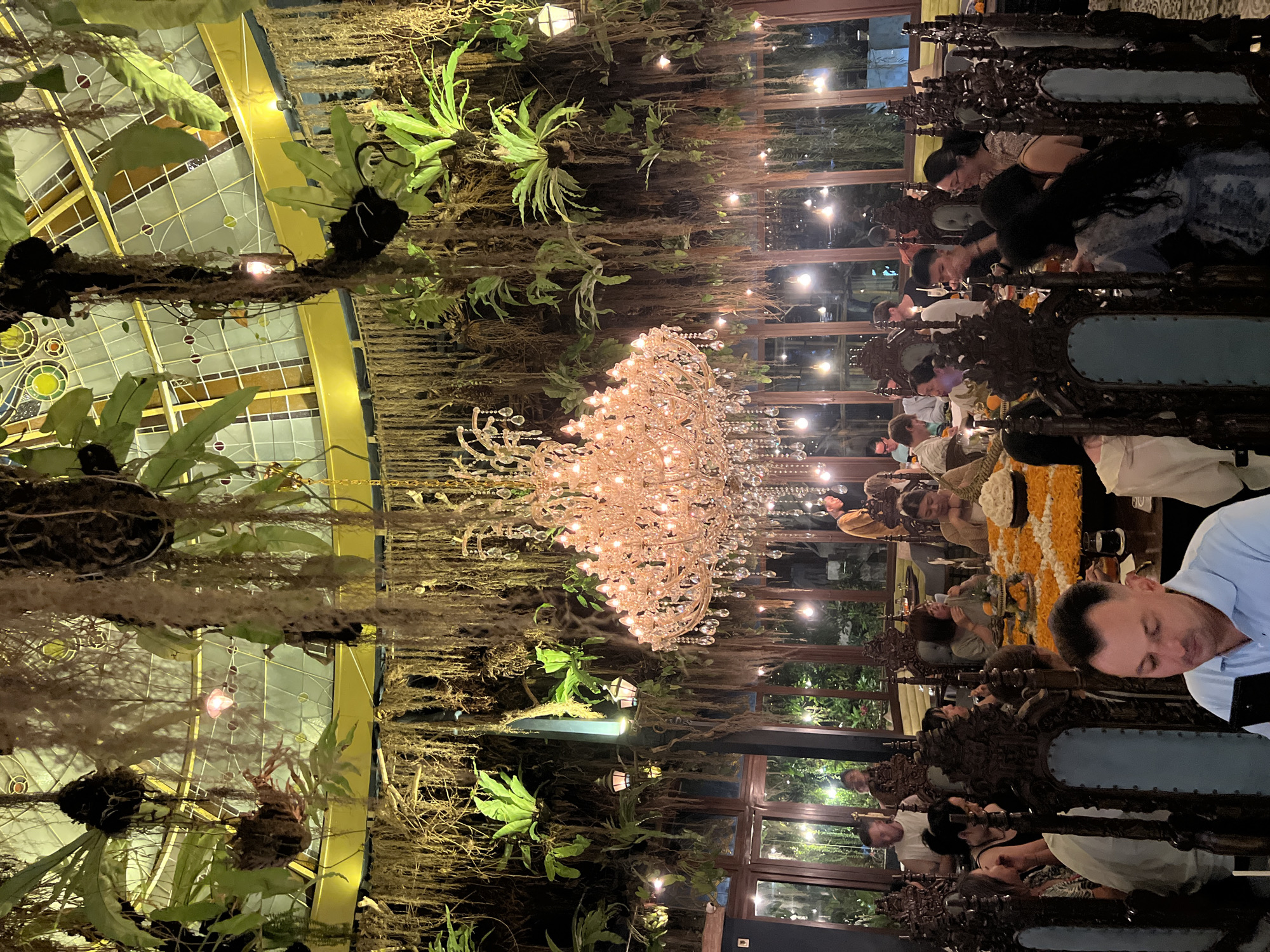 The image depicts a dining scene at Locavore, located in the Ubud District of Indonesia. Multiple individuals are seated at ornately carved chairs, appearing to be engaged in dining or taking photos of the table setting. The table is adorned with an orange and yellow floral arrangement, possibly marigolds. Overhead, a large, multi-tiered crystal chandelier dominates the view, surrounded by hanging plants, including ferns and Spanish moss. The ceiling features a stained-glass section, suggesting a greenhouse or conservatory setting. Tall windows line the perimeter of the room, offering glimpses of foliage outside. The lighting is soft and warm, creating an intimate and upscale dining atmosphere.