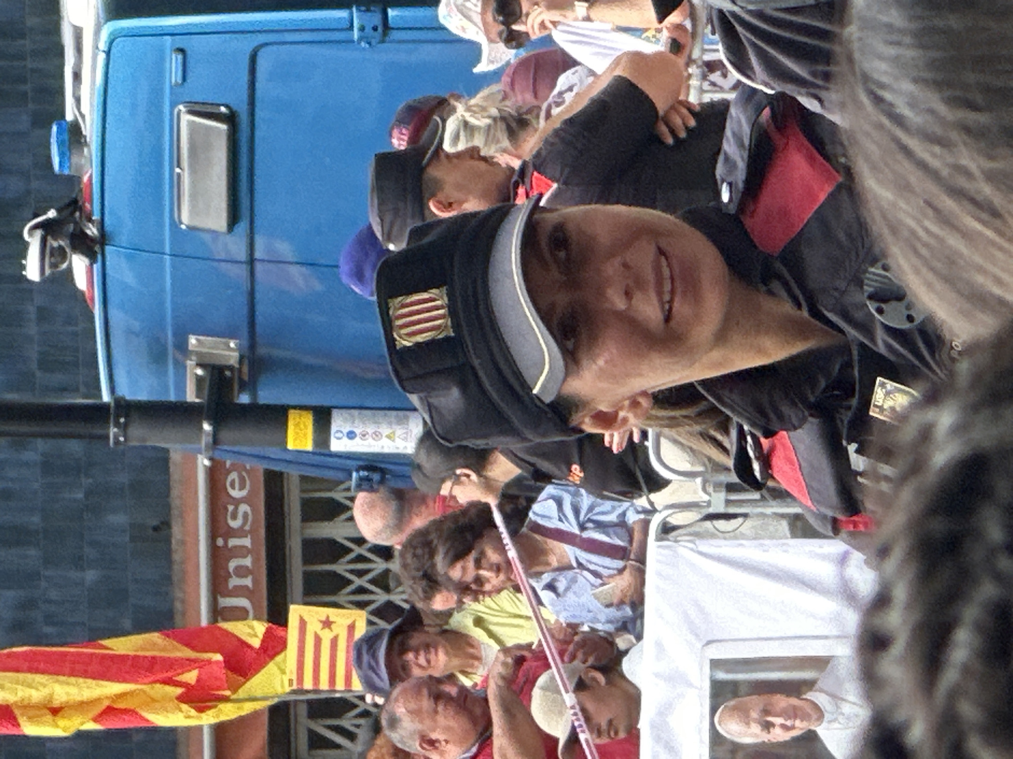 In an outdoor urban setting, likely a public gathering, a group of people are visible. A blue vehicle, possibly a van or truck, is prominent in the background, with a security camera mounted on its roof. A flag with red and yellow stripes is displayed on a pole. Several individuals are wearing uniforms, including caps with emblems. One person in the foreground is wearing a dark uniform with red accents and a cap featuring a coat of arms. Another individual in the foreground, possibly a woman, is looking towards the camera with a slight smile. A framed photograph of a man in religious attire is visible to the left. The environment appears to be a street or plaza, with barriers and a crowd of onlookers suggesting an event or demonstration.
