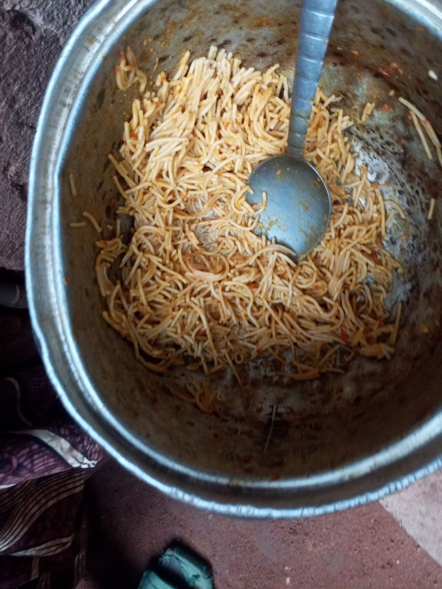 In Tempure, Nigeria, a metal pot filled with spaghetti is visible. A metal ladle rests inside the pot, partially submerged in the pasta. The spaghetti appears to be cooked and coated in a reddish sauce, possibly tomato-based. The pot is set on a reddish-brown surface, possibly brick or stone, with a patterned fabric visible to the left and a rough, textured surface above the pot. The overall scene suggests a casual, perhaps outdoor, setting where food is being prepared or served. There are no people or animals visible in the image.
