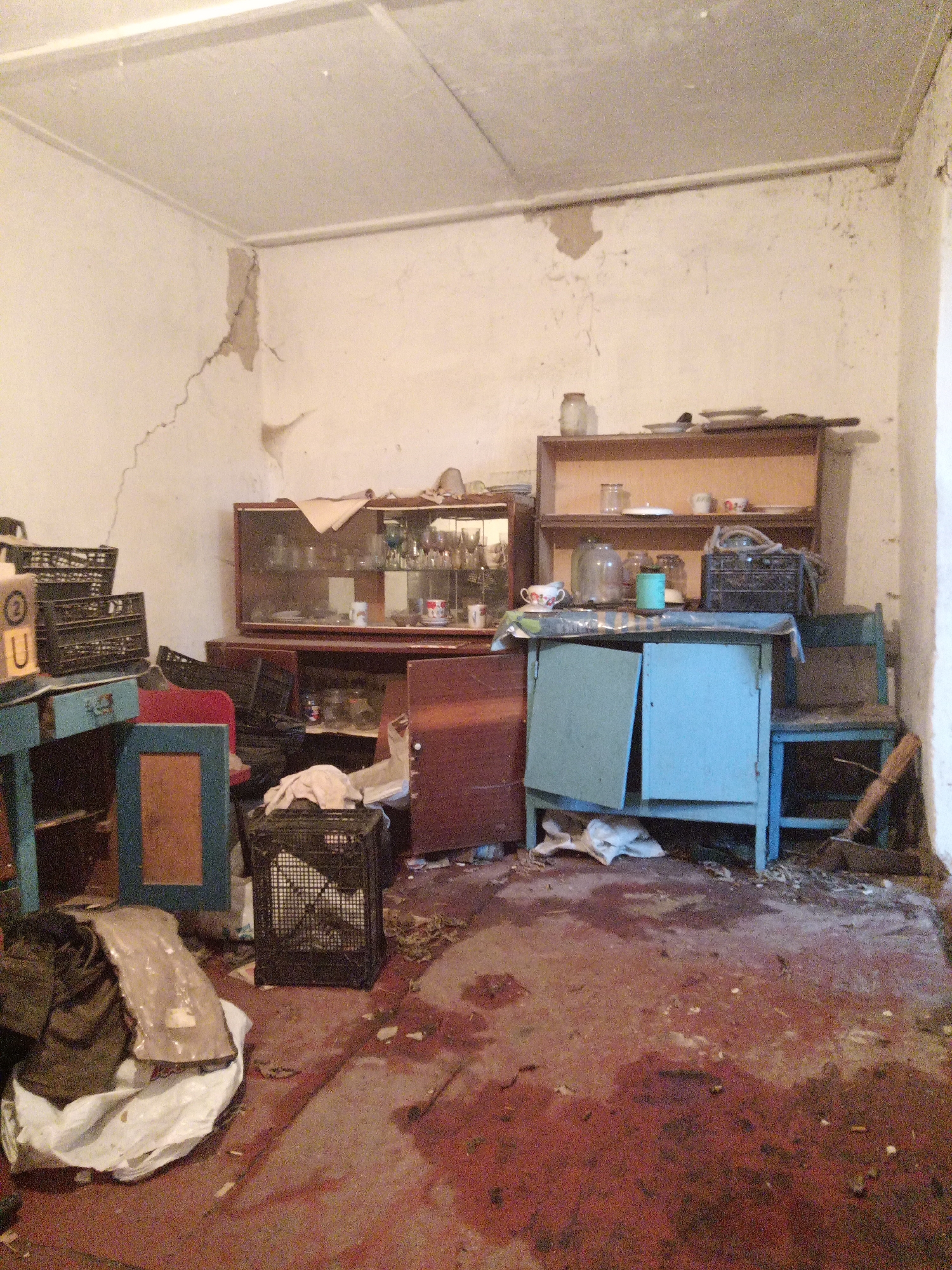The image depicts an interior scene, likely a room in a dwelling, in Veremia, Ukraine. The environment appears neglected and shows signs of disrepair. The walls are a light color, possibly white or off-white, and exhibit significant cracking, particularly in the upper left corner. A portion of the ceiling is visible, also showing signs of wear and staining.
The room is furnished with several pieces of furniture. A large, dark-colored cabinet with glass doors is positioned against the back wall, containing an assortment of glassware and some dishes. Above this cabinet, a wooden shelf unit holds more glassware, dishes, and a jar. In front of the cabinet is a light blue, wooden piece of furniture, possibly a small desk or cabinet, with its doors ajar. To the left of this blue furniture, a smaller, darker blue wooden desk or table is visible, with stacked black plastic crates on top. To the right of the blue furniture, a simple blue wooden chair or bench is situated.
Various objects are scattered around the room, contributing to the appearance of disarray. Black plastic crates are present in multiple locations. A dark-colored bag or bundle of clothing is on the floor in the lower left corner, next to a crumpled white sheet or tarp. The floor itself is a dark, reddish-brown color and appears stained or dirty, with scattered debris and what might be dried liquid stains. In the lower right corner, a broom is leaning against the wall.
The overall impression is one of abandonment or severe neglect, suggesting a dwelling that has fallen into disuse or is in a state of disrepair. There are no visible individuals present in the image.