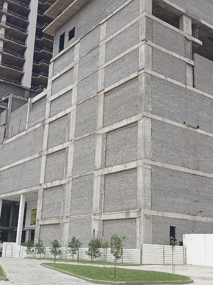 The image captures an exterior view of a large, multi-story building under construction, dominated by gray concrete and blockwork. The building in the foreground shows at least five visible stories, characterized by a grid-like facade of reinforced concrete columns and beams, with the spaces between filled by light gray cinder blocks laid in a stretcher bond pattern. The building presents two sides, meeting at a prominent outer corner towards the right of the frame.
On the upper left side of the main building, two small, dark rectangular openings are visible, possibly future windows or service vents. On the ground floor, on the left side, there is a wider entrance area with a white column and a yellow sign featuring unreadable text set further back inside. On the right side of the building's ground floor, a single black door is visible against the block wall.
Behind this main structure, a taller, narrower building is also under construction, with exposed concrete slabs defining its multiple floors and visible rebar or structural elements.
In the foreground, a landscaped area runs along the base of the main building. This area features a strip of green grass with several small, newly planted trees spaced regularly along it. A low, white fence with horizontal slats separates this grassy strip from a paved walkway or driveway made of light gray pavers, which curves from the bottom left of the image.
The scene depicts an active construction site, though no workers or heavy machinery are visible. The time of day appears to be daytime, with soft, even lighting suggesting overcast conditions or diffuse sunlight, as there are no harsh shadows. The overall color palette is dominated by grays from the construction materials, accented by the green of the grass and trees, and the white of the fence and column. No specific location or emotions are discernible.