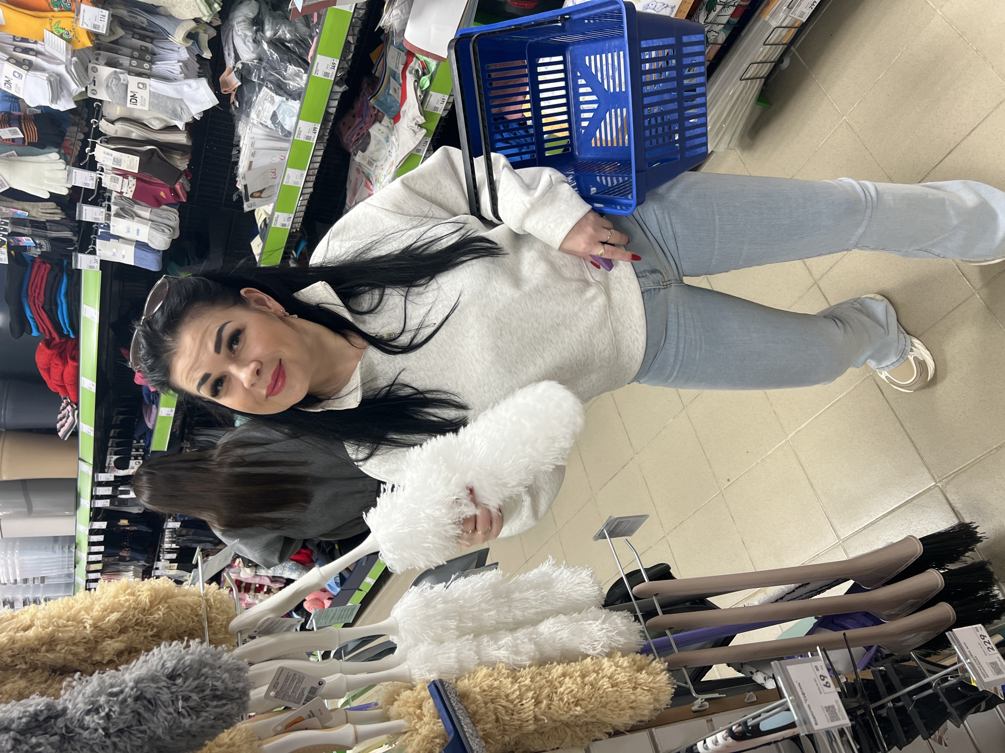 A woman is standing in a store aisle, holding a white duster in her right hand, while holding a blue shopping basket with her left. She is wearing a light-grey sweatshirt, blue jeans, and white sneakers. She has long, dark hair and is looking at the camera. She has a pair of sunglasses resting on her head. The store aisle contains shelves with socks, dusters, and other items. Price tags are visible on the shelves. The location is the city of Chaikovsky, Russia.