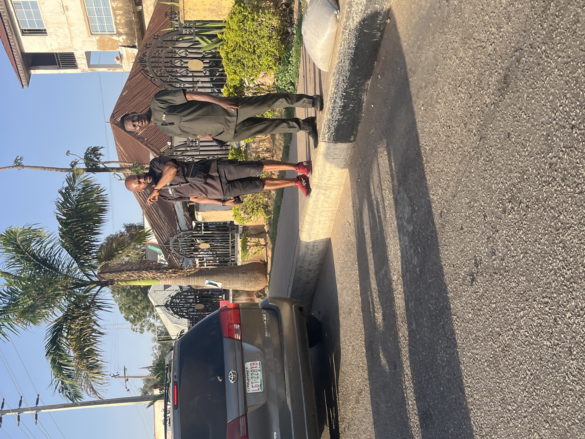 Two men stand on a sidewalk next to a street in Gura Top, Nigeria. One man wears a dark green uniform. The other man wears a black shirt, black shorts, and red sneakers. A silver minivan with license plate LGT-228XB is parked on the side of the street. A palm tree is visible. A building is visible behind the men.