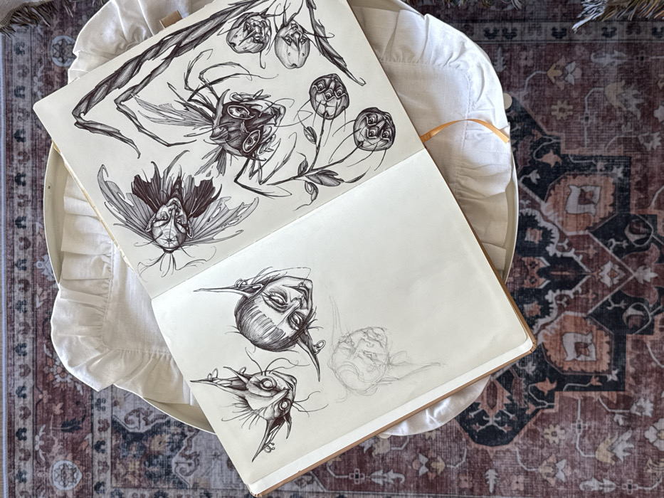 An open, brown-bound sketchbook lies centered on a light cream-colored, ruffled fabric surface, positioned above a multi-patterned rug. The image is captured from a top-down perspective indoors.
The sketchbook's left page features two distinct stylized ink drawings of creature heads with elongated, pointed ears. The upper drawing depicts a wide-eyed, grinning head with long eyelashes and fine facial hair. The lower, more centered drawing shows a head with bangs, prominent pointed ears adorned with small rings, and a subtle smirk. Below these, a faint pencil sketch of another head is visible.
The sketchbook's right page presents a collection of more elaborate ink drawings. A winged creature with a stylized head, multiple eyes, and sharp features dominates the upper-left. Below and to its right, a larger, intricate insect-like creature with numerous legs and eyes is depicted. This creature is integrated with leafy branches that extend downwards, bearing several cherry-like fruits, each meticulously drawn to feature multiple eyes and distinct facial expressions. A thin orange ribbon bookmark extends from beneath the right page.
The cream-colored ruffled fabric beneath the sketchbook appears to be part of a round furnishing, possibly a stool or cushioned tray. Below this furnishing, a woven rug covers the floor, displaying an ornate, complex pattern primarily in shades of dark red, burgundy, dark blue, beige, and dark brown. The pattern includes intricate floral and geometric motifs.
No identifiable architectural features, landmarks, or unique cultural elements are present to determine a specific location within the city of Moscow, Russia.